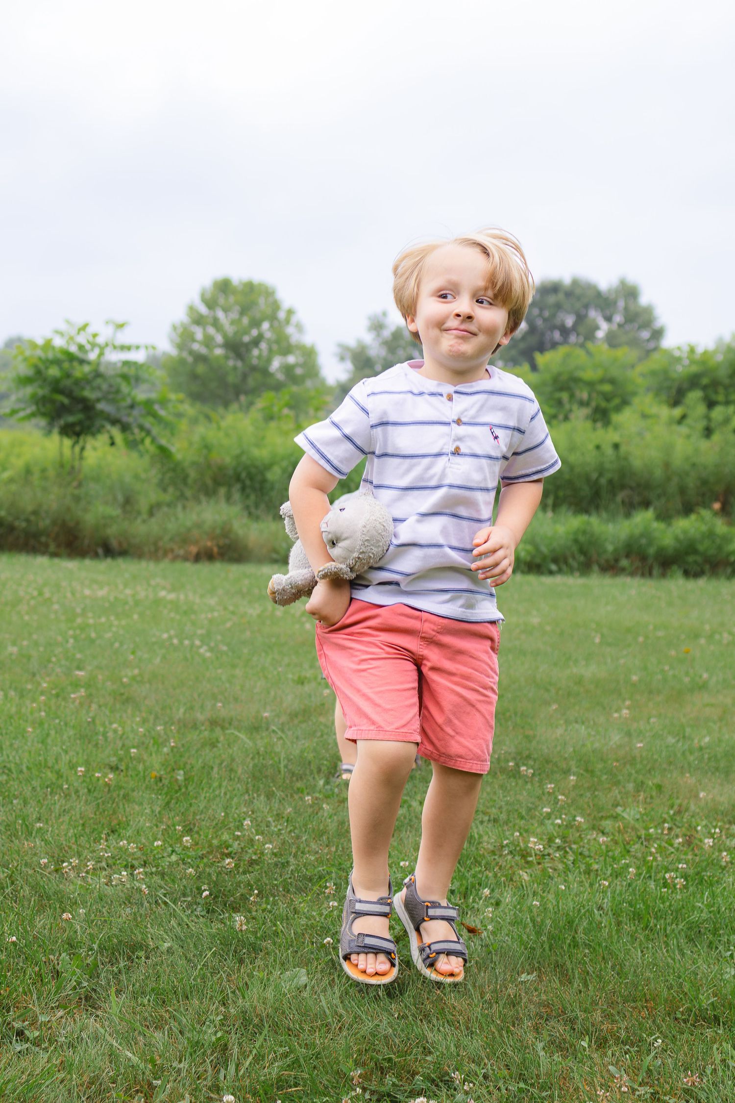 Cute blonde boy in striped shirt holding a stuffed animal toy in a green park.