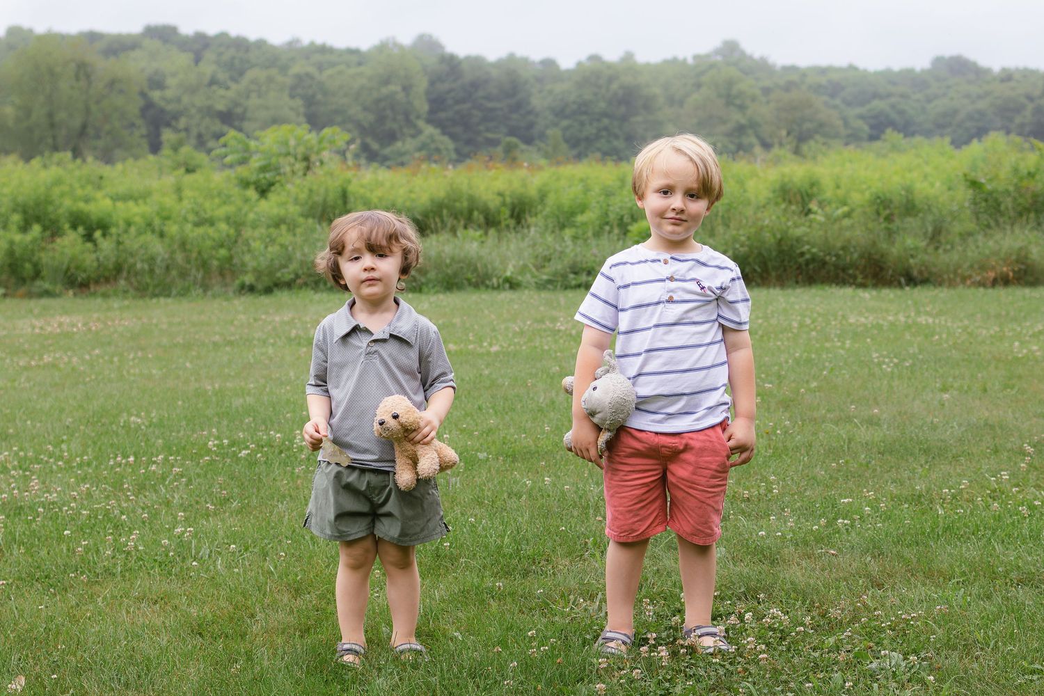 Two young boys stand in a grassy field holding stuffed animals, posing for an outdoor portrait.
