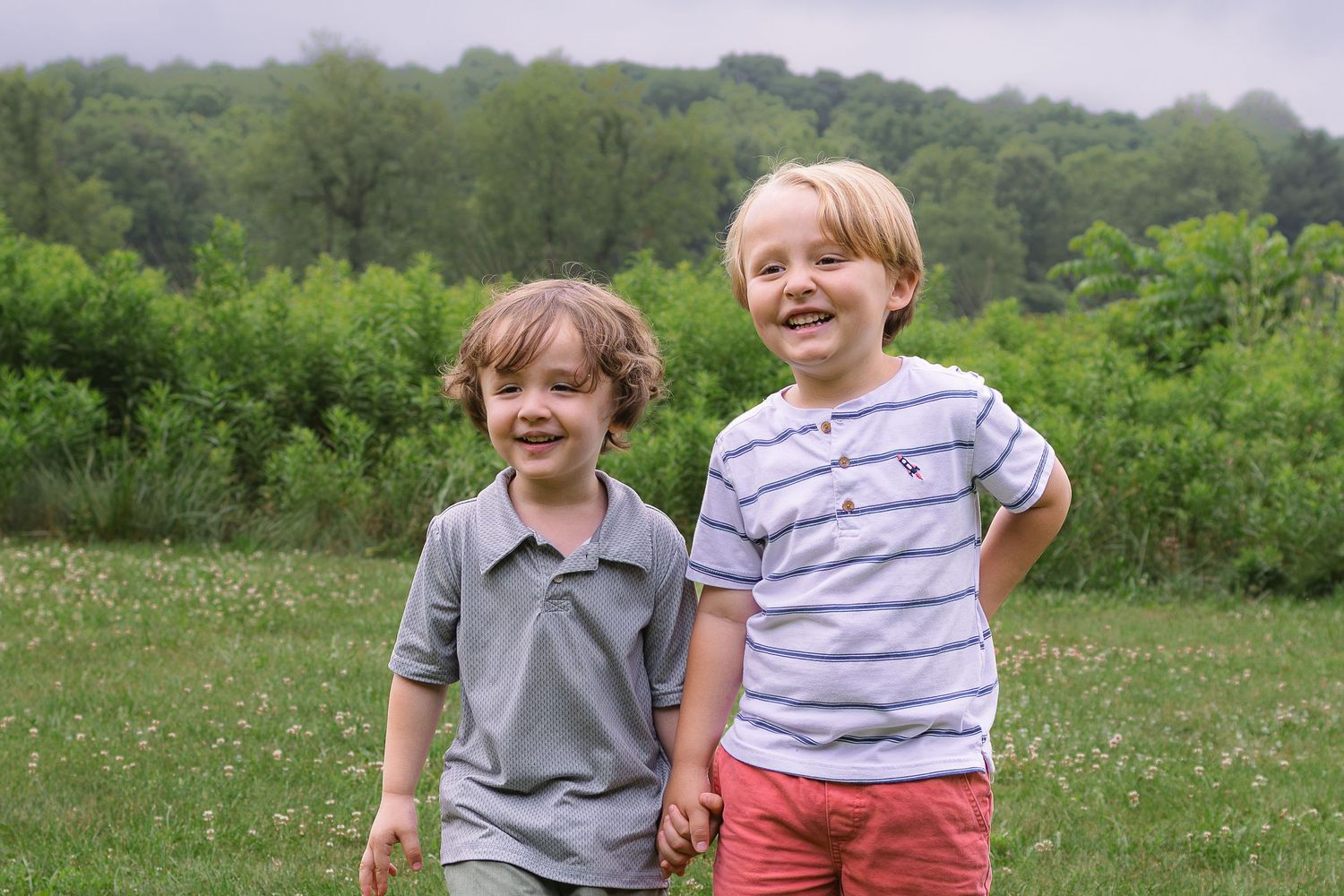 Two happy young boys holding hands and smiling in a green grassy field with trees in the background.