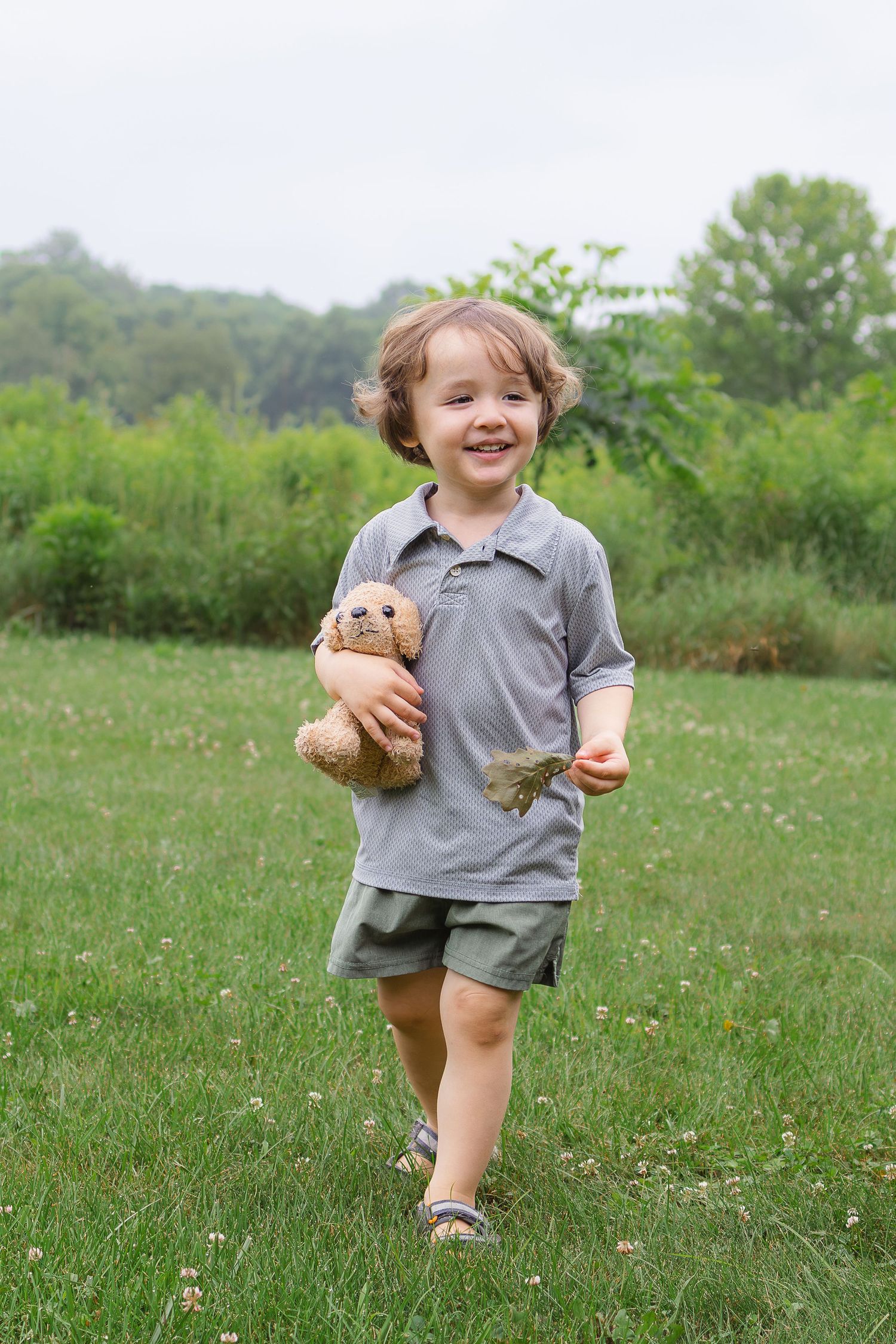A happy young boy runs through a green grassy field while holding a brown stuffed animal dog toy.