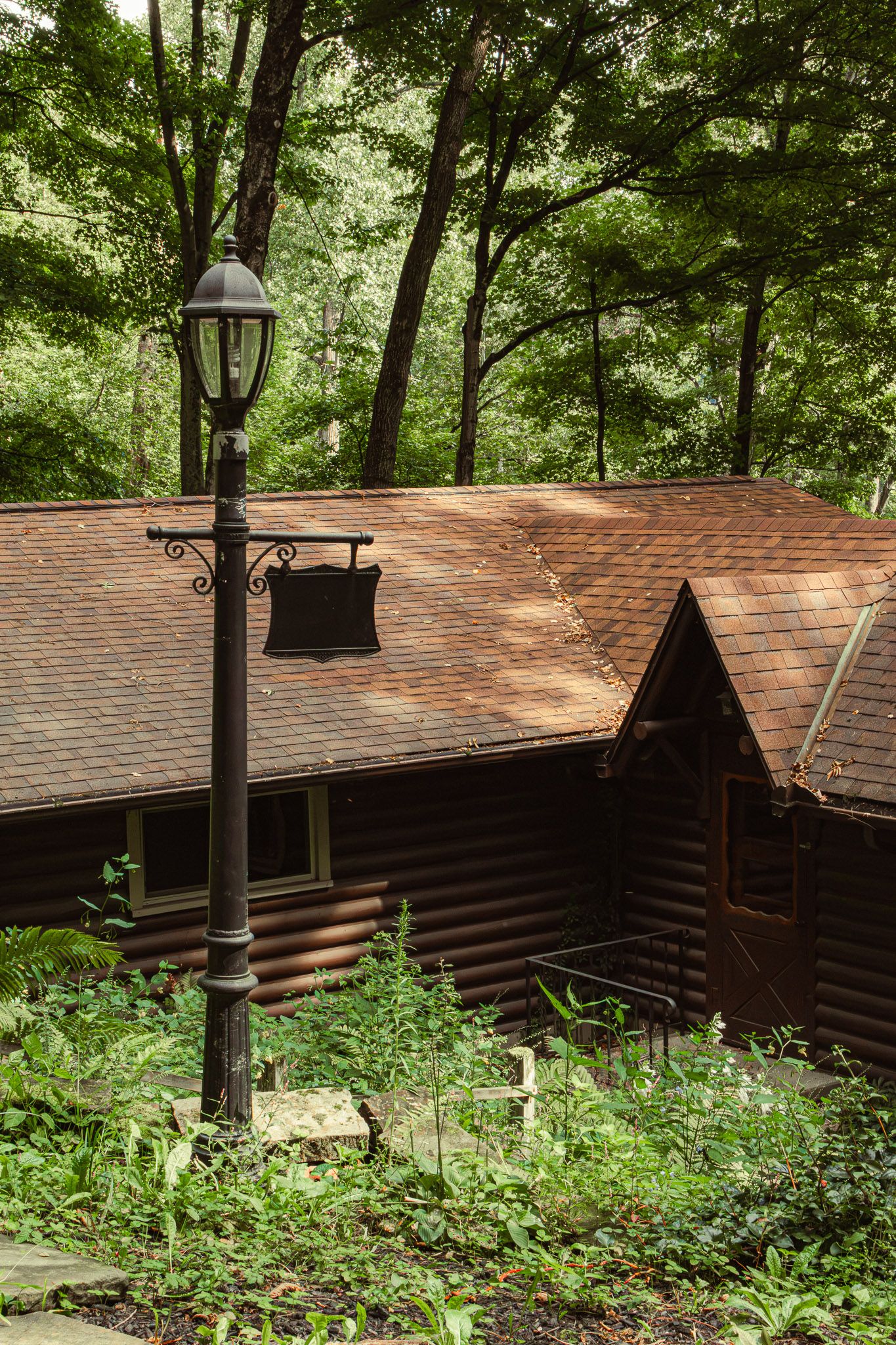 Rustic log cabin with brown shingle roof surrounded by lush green forest trees and a vintage black lamp post in foreground.