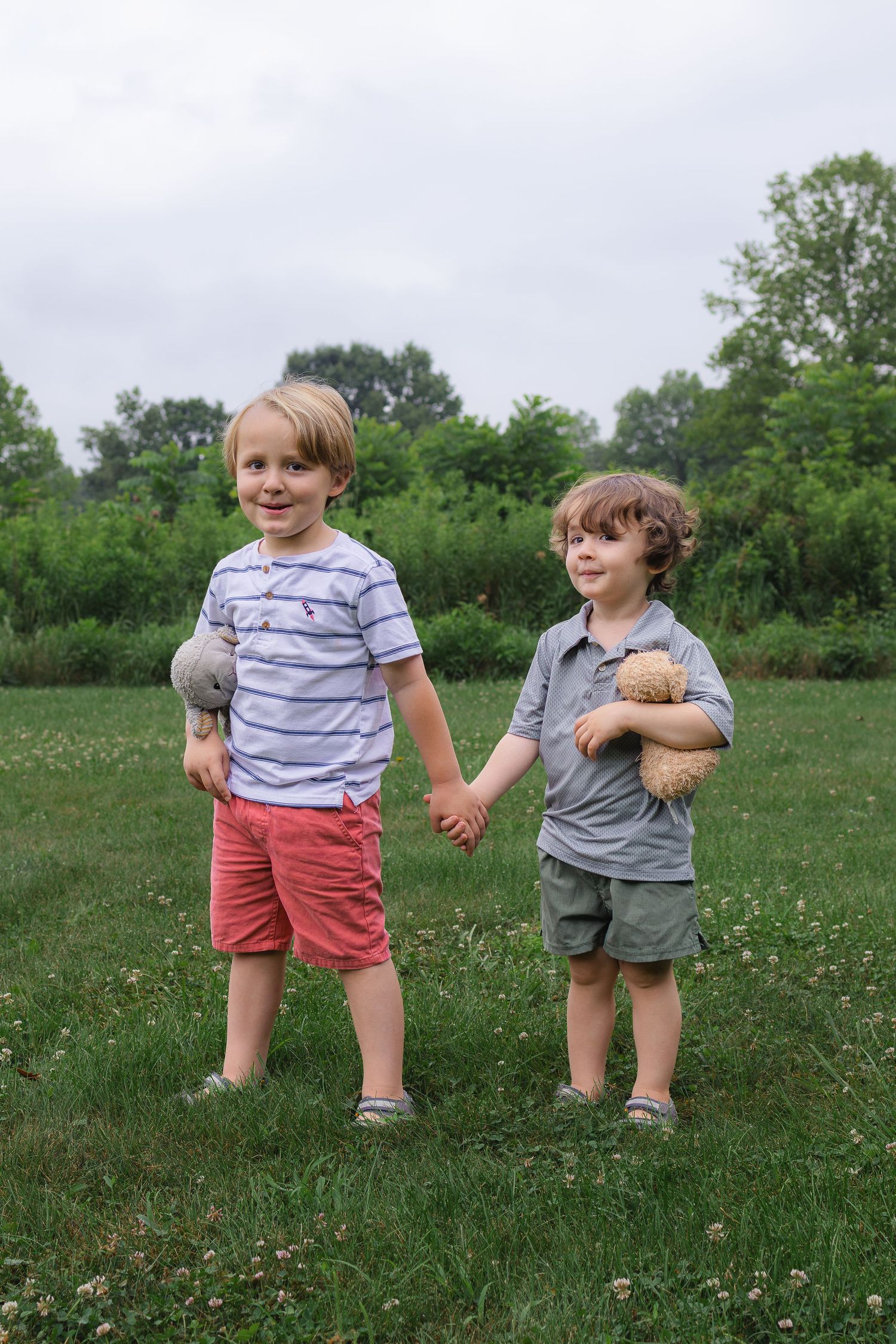 Two young boys holding hands in a green field, one holding a stuffed animal and the other a toy.