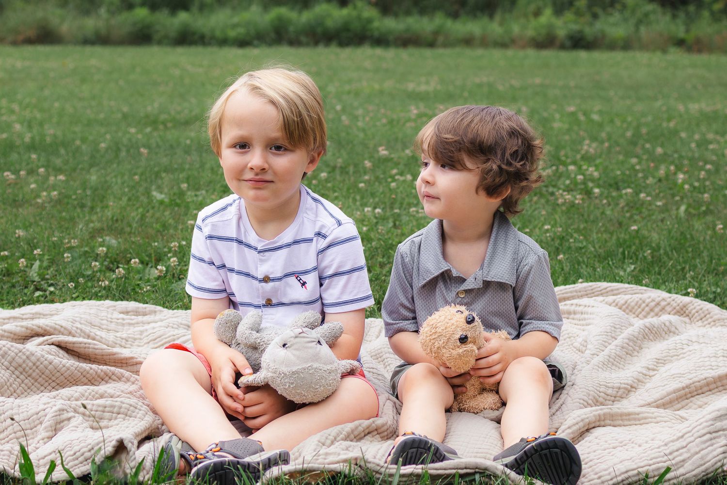 Two young boys sit on a blanket in a field, each holding a stuffed animal, looking away.