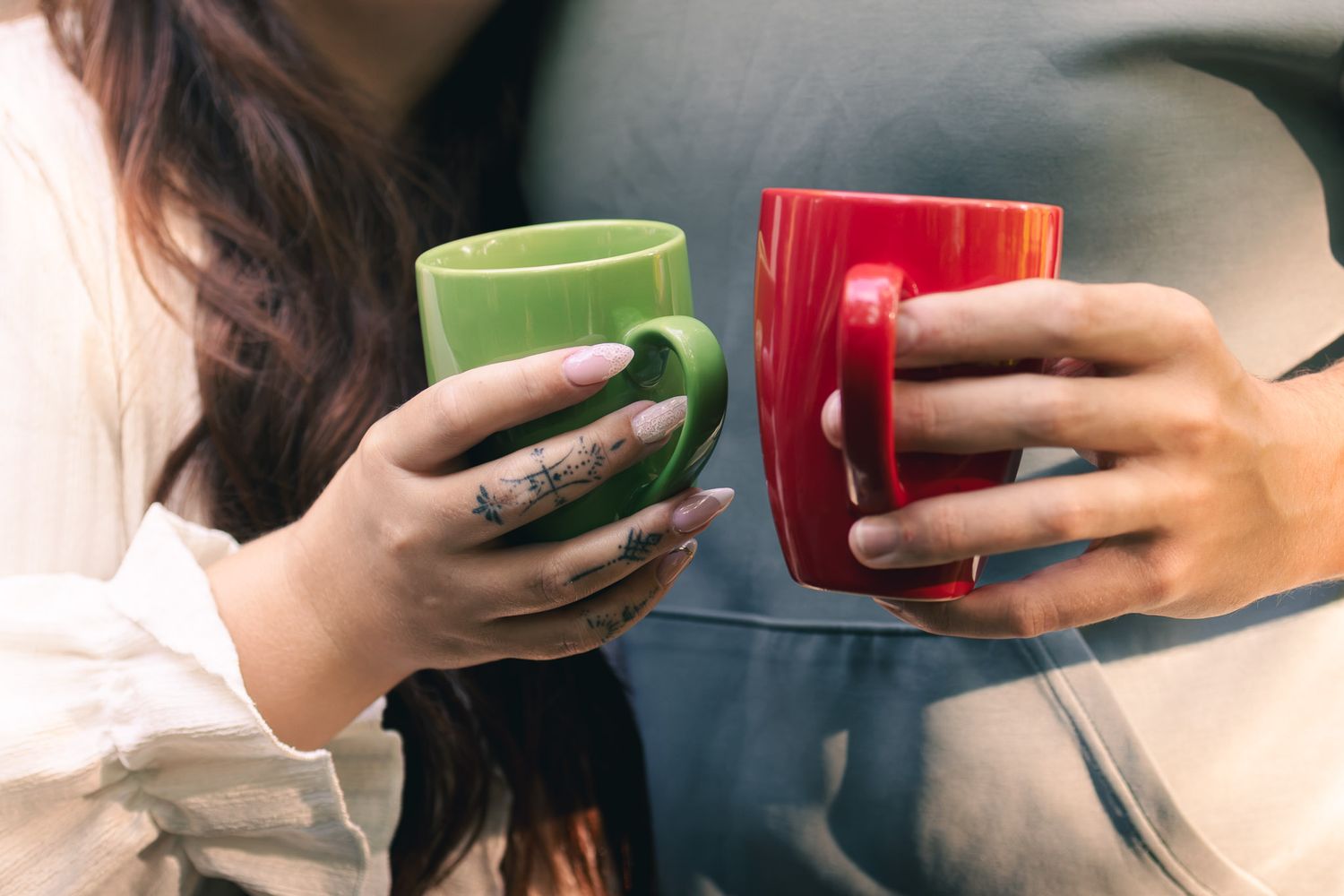 Two women holding green and red ceramic mugs together, with tattooed hands visible, sitting on a couch.
