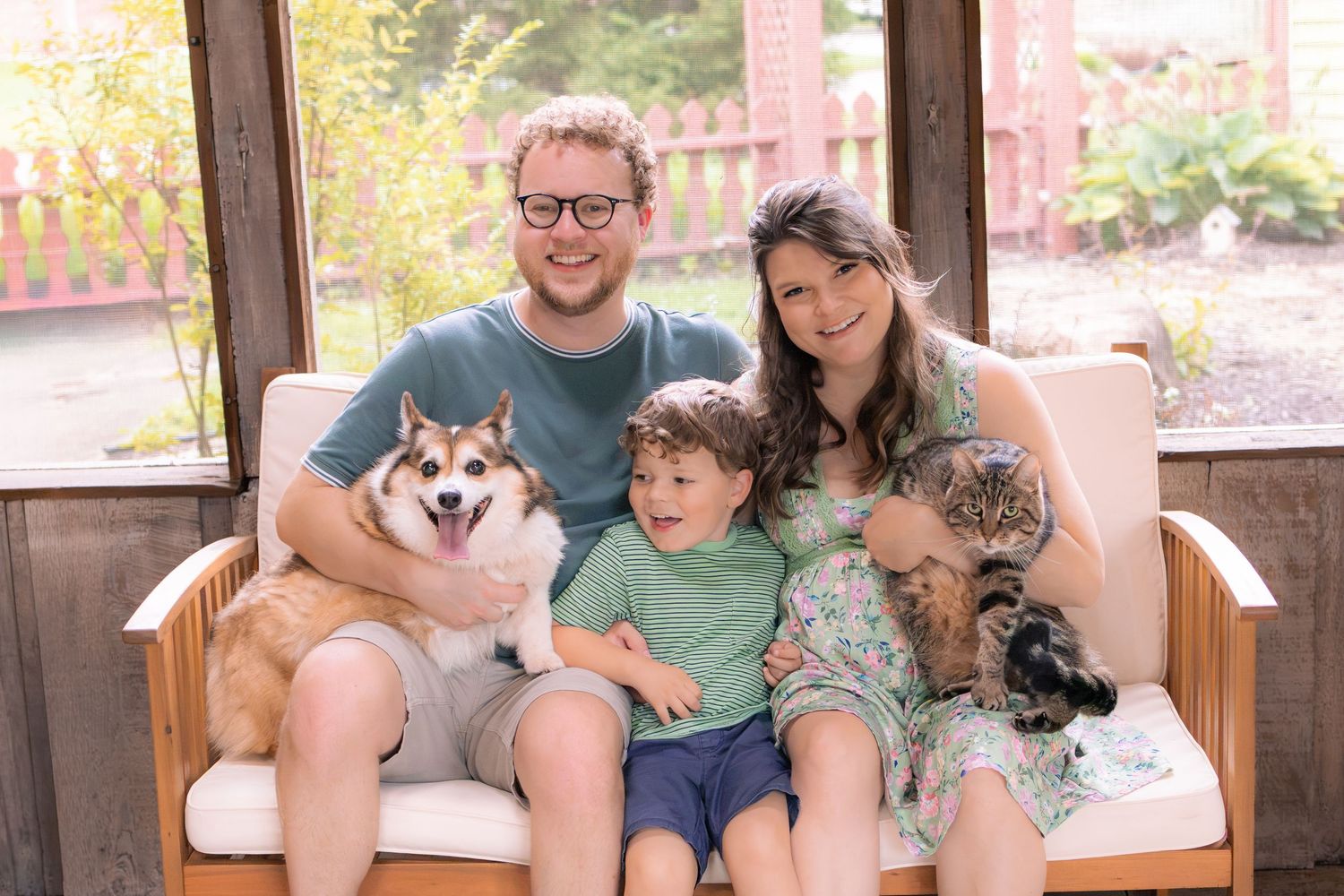 A happy family portrait with parents, young child, a husky dog, and a cat on a porch.