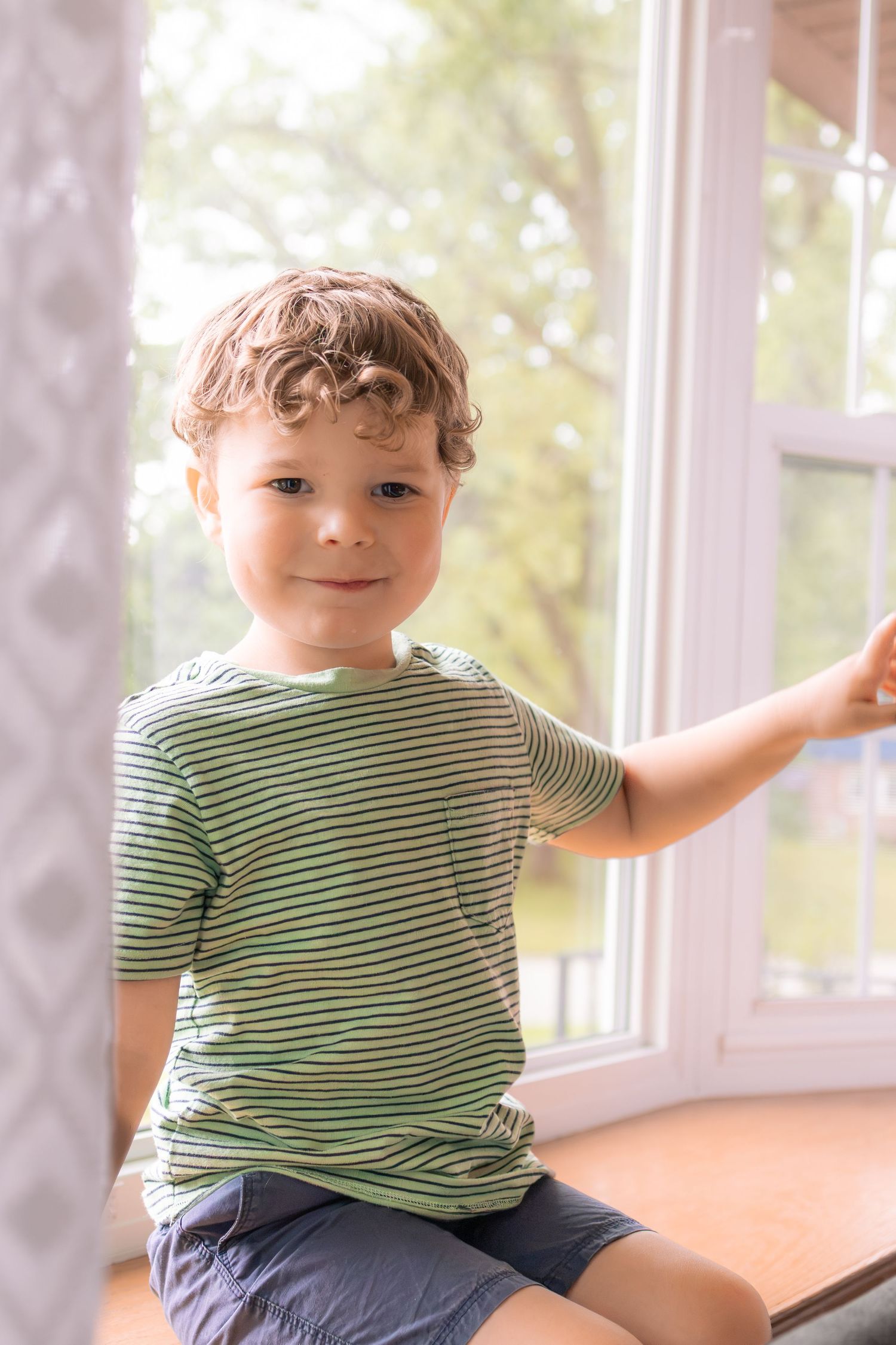Smiling young boy in striped shirt sitting on windowsill by bright window.