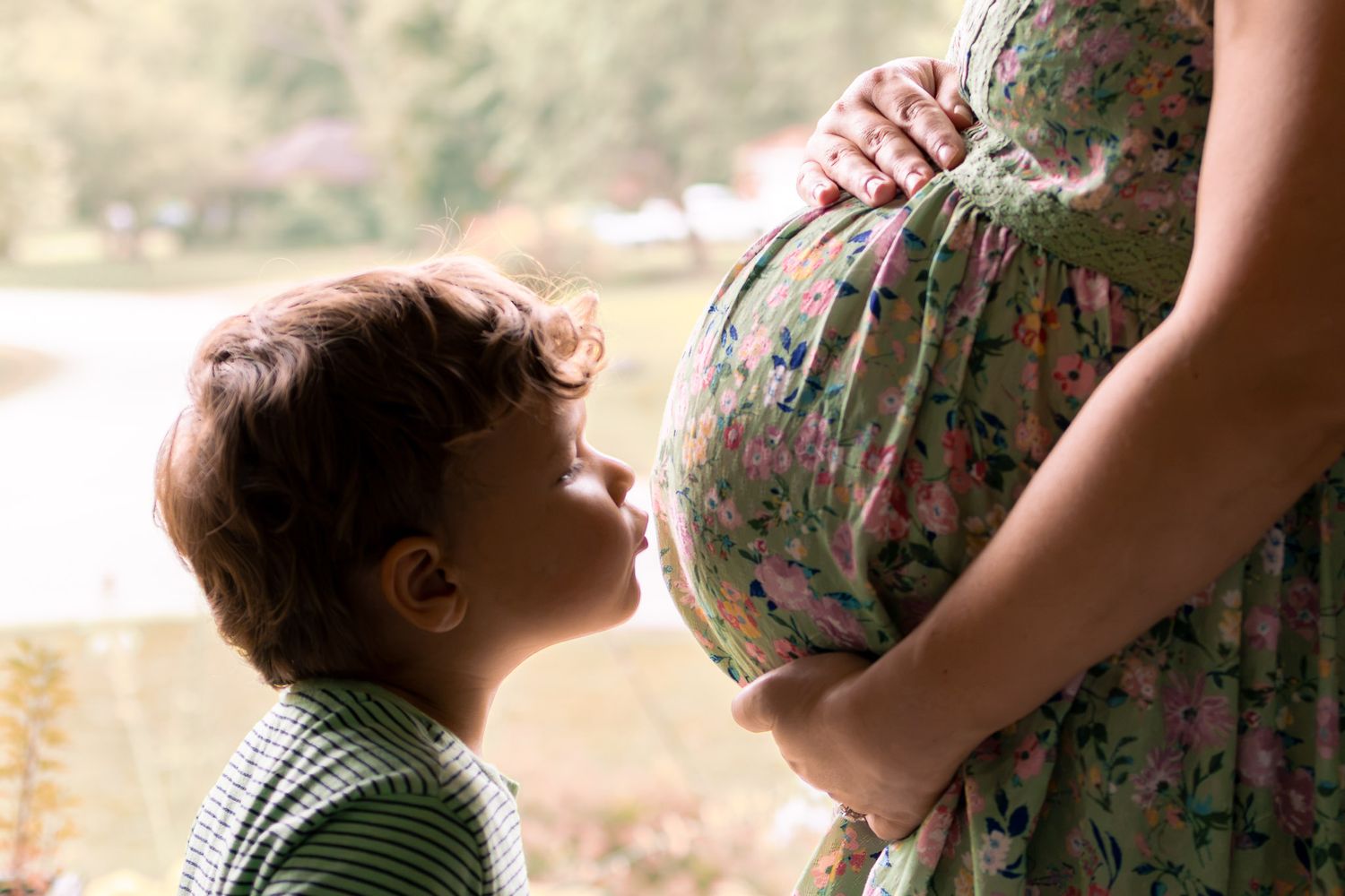 Young boy kissing pregnant mother's belly in a tender maternity moment near a sunny window.
