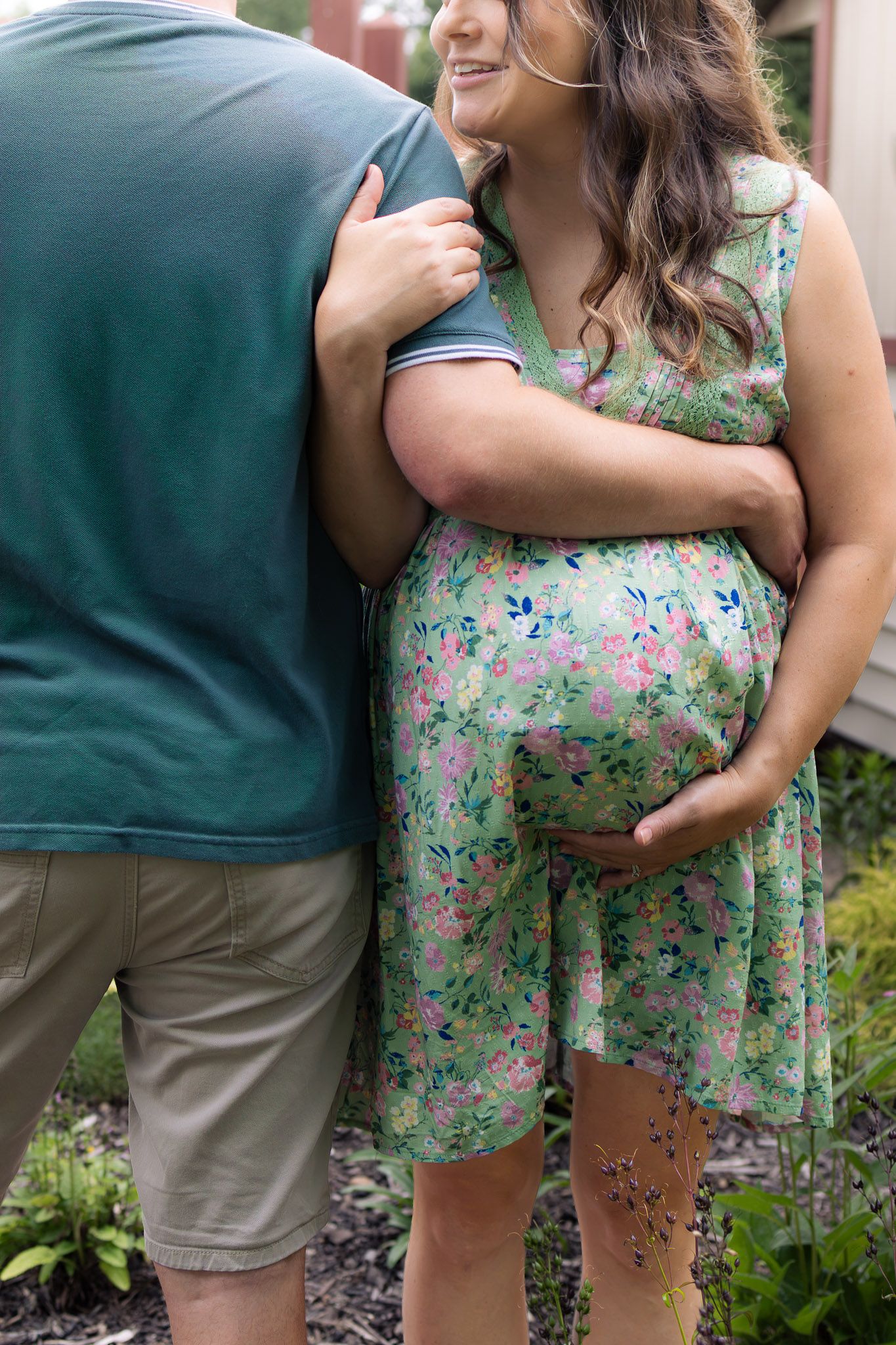 A smiling pregnant woman in a green floral dress embraces her partner in a garden.