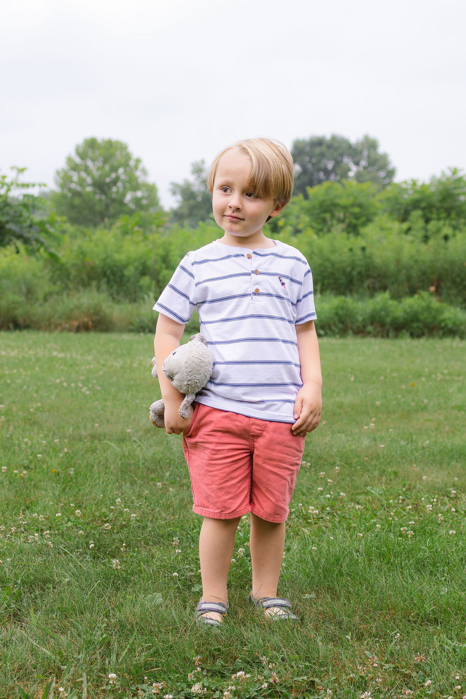 A young blonde boy stands on a green grassy field holding a small gray stuffed animal toy.