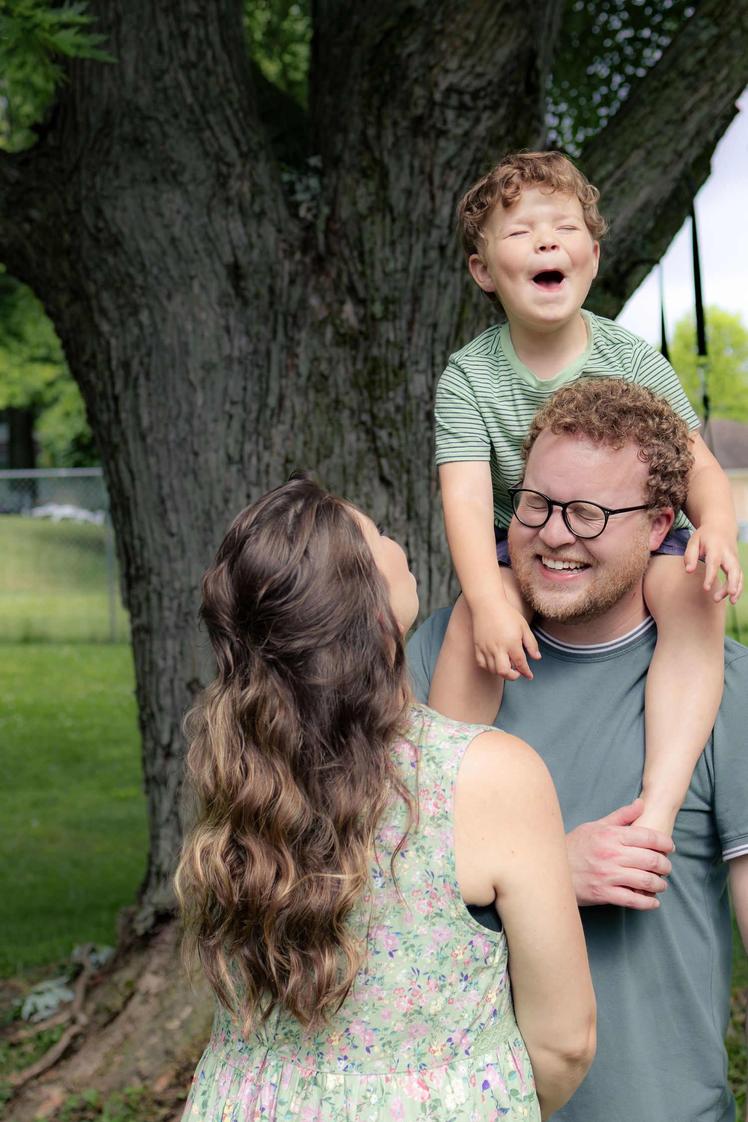 Laughing young boy sitting on dad's shoulders while mom reaches up in a park setting.