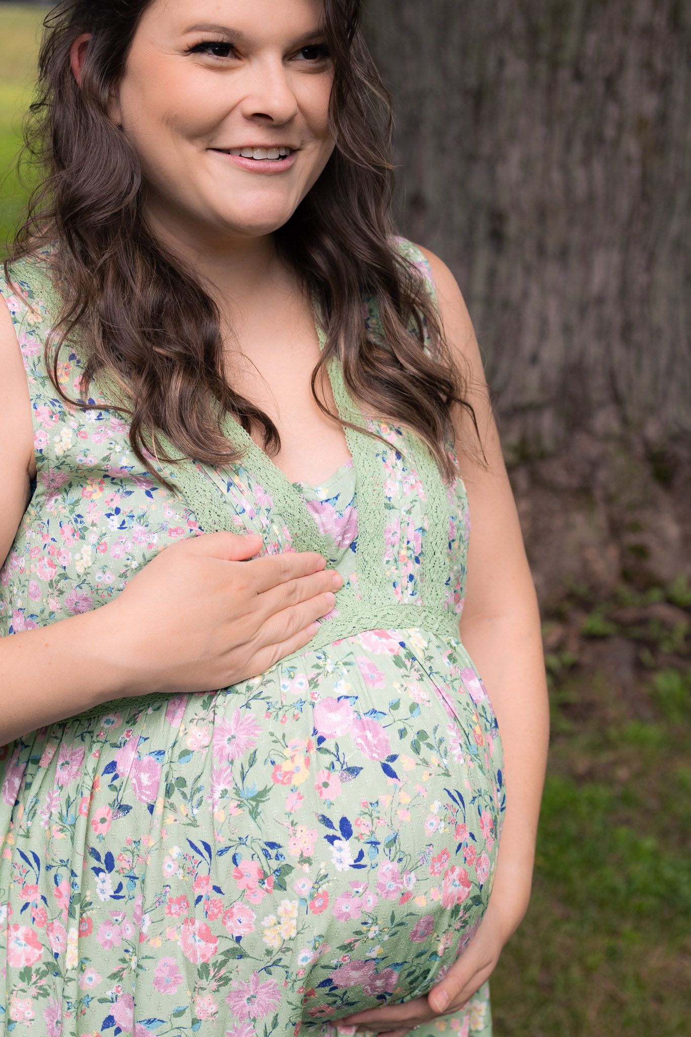 Happy pregnant woman in green floral dress holding baby bump outdoors near tree.