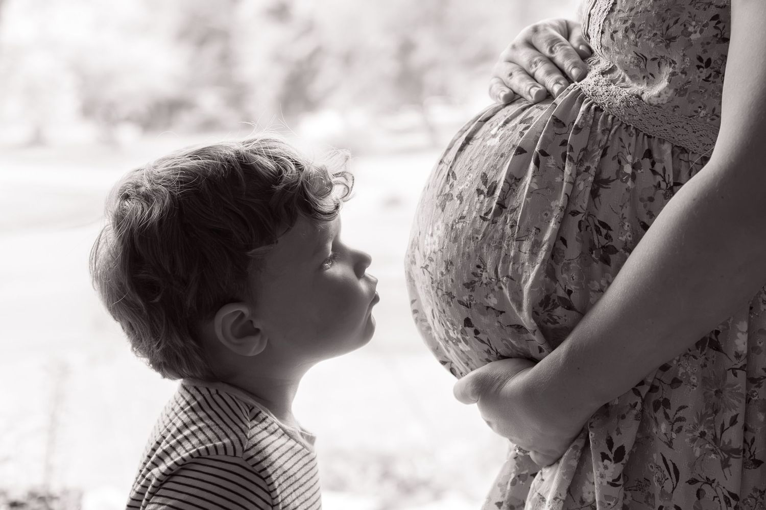 Black and white photo of young boy looking up at pregnant mothers belly lovingly.