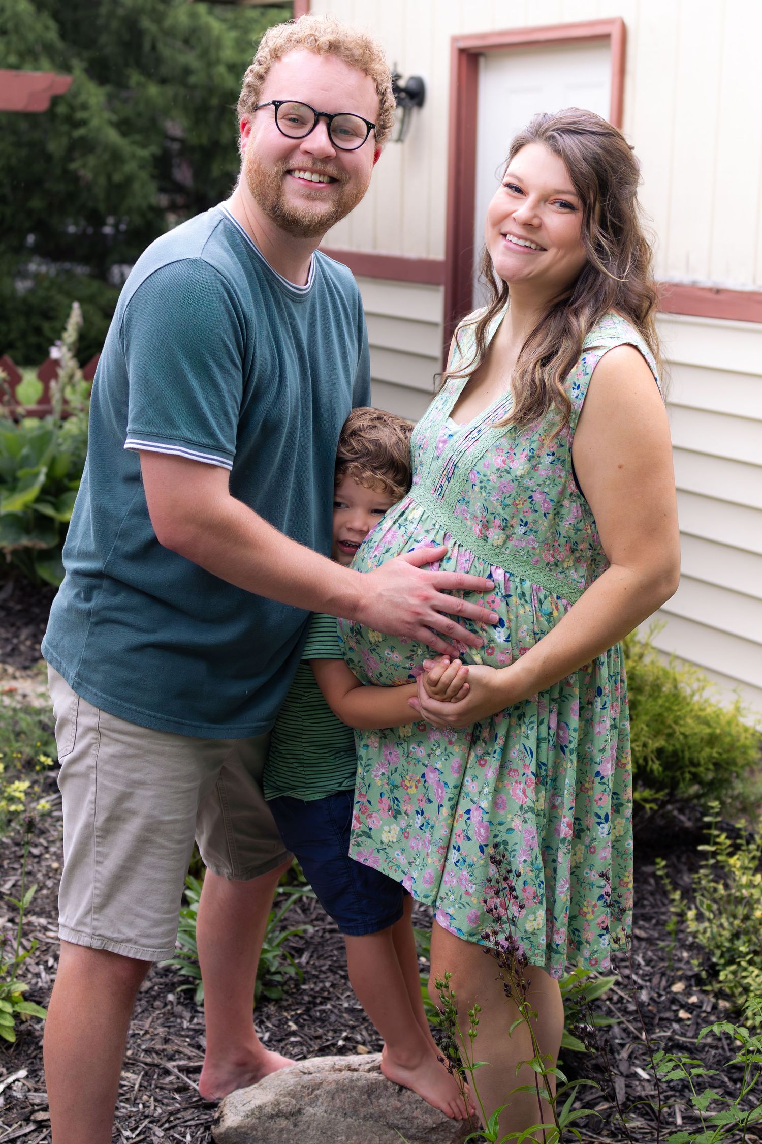 Happy family with young child posing outdoors with pregnant mother in floral dress.