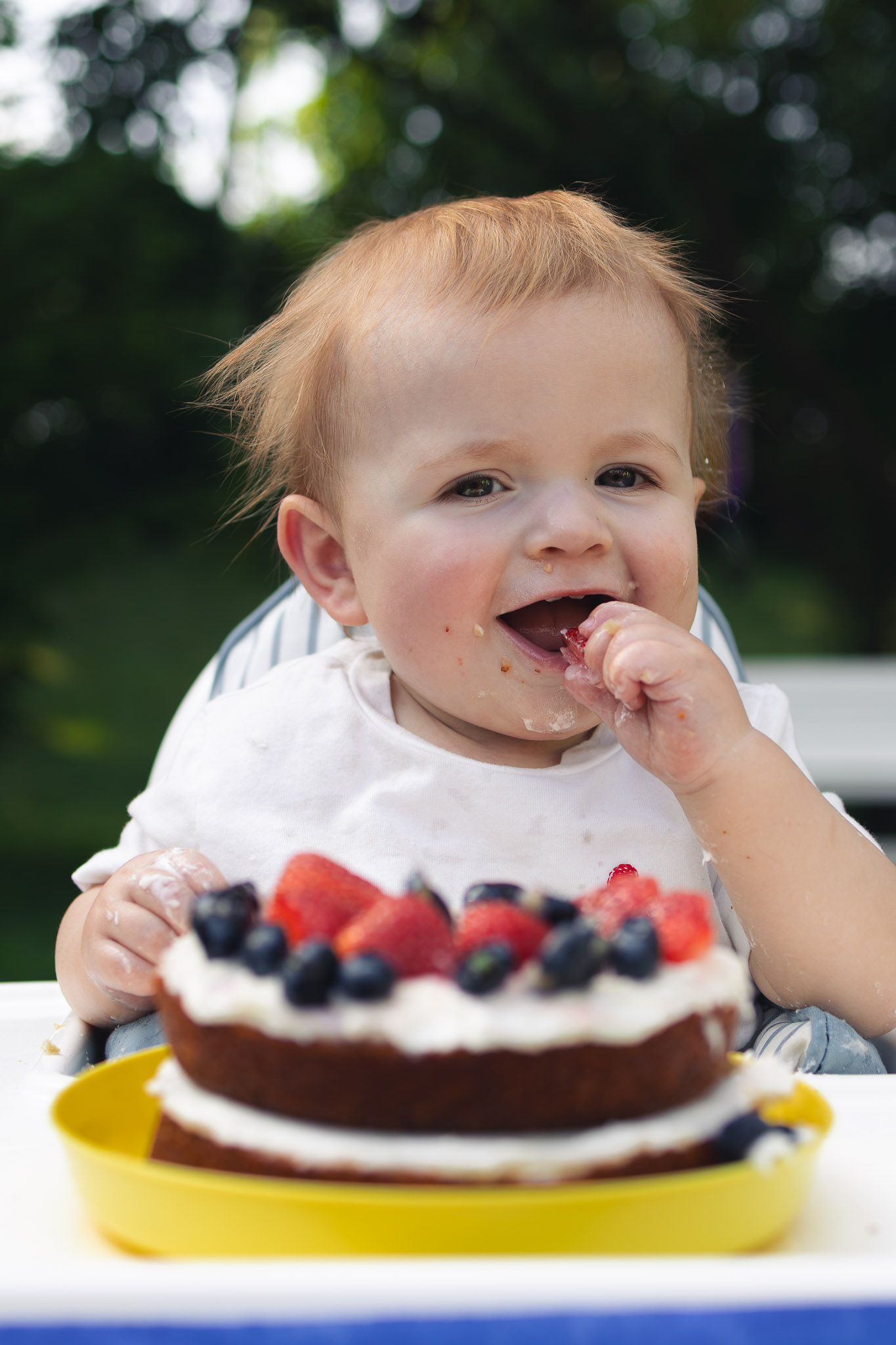 Smiling baby in a high chair enjoying a first birthday smash cake topped with strawberries and blueberries on a yellow plate.