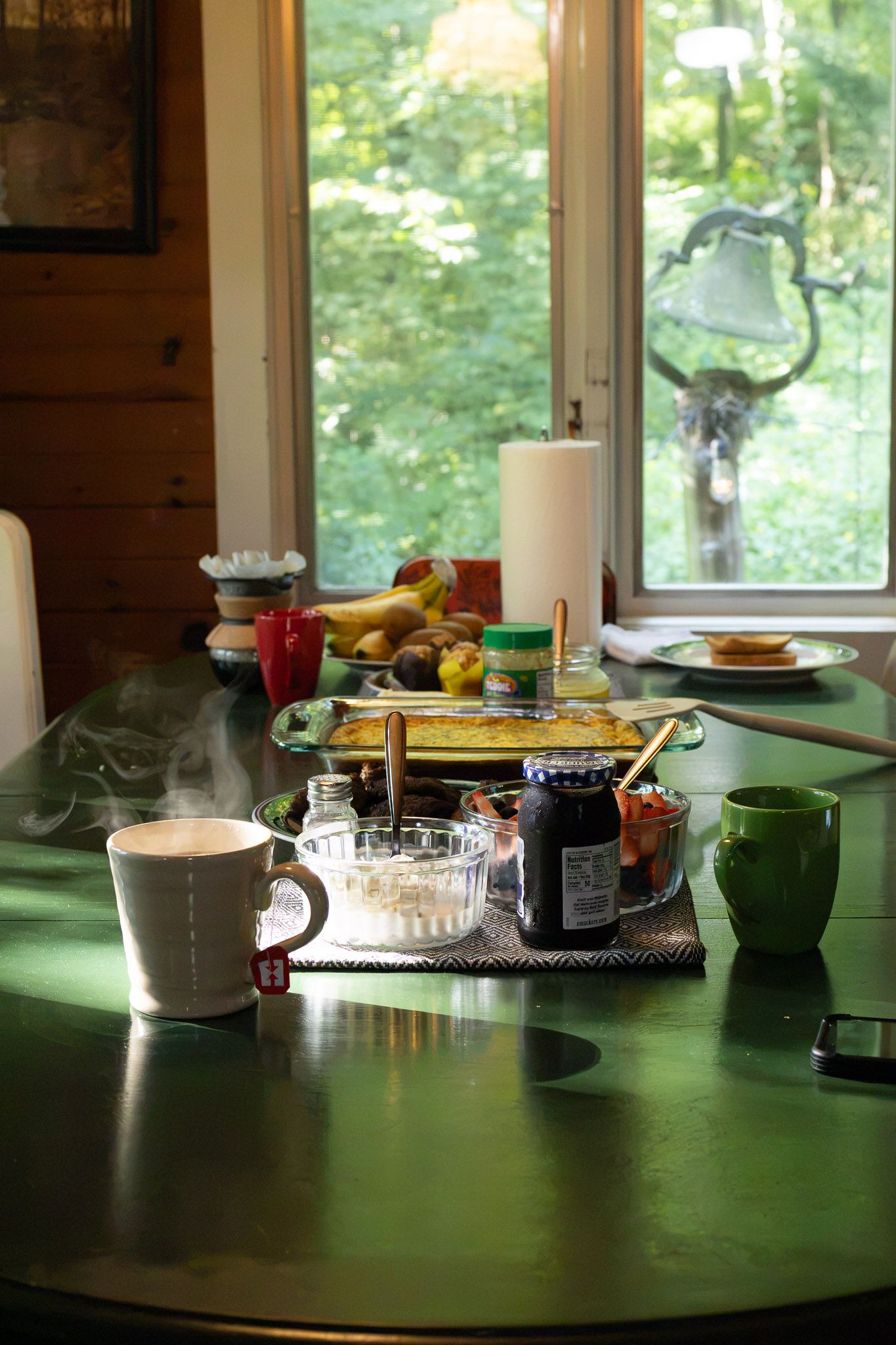 Cozy cabin breakfast table with steaming coffee, jam, fruit, and baked goods near a window overlooking lush green trees.