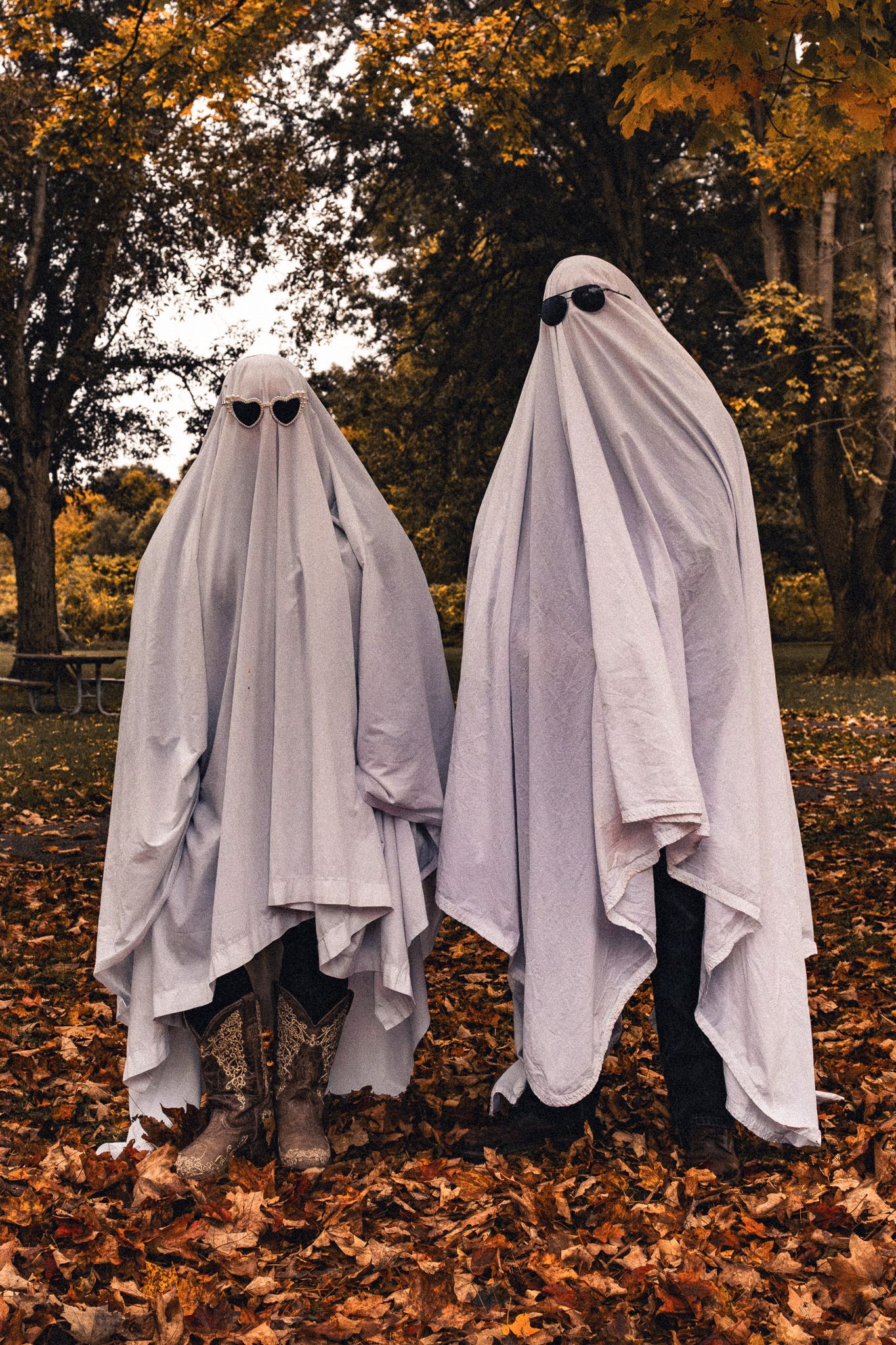 Two people in classic white ghost costumes with black eye holes stand among fallen autumn leaves in a park.