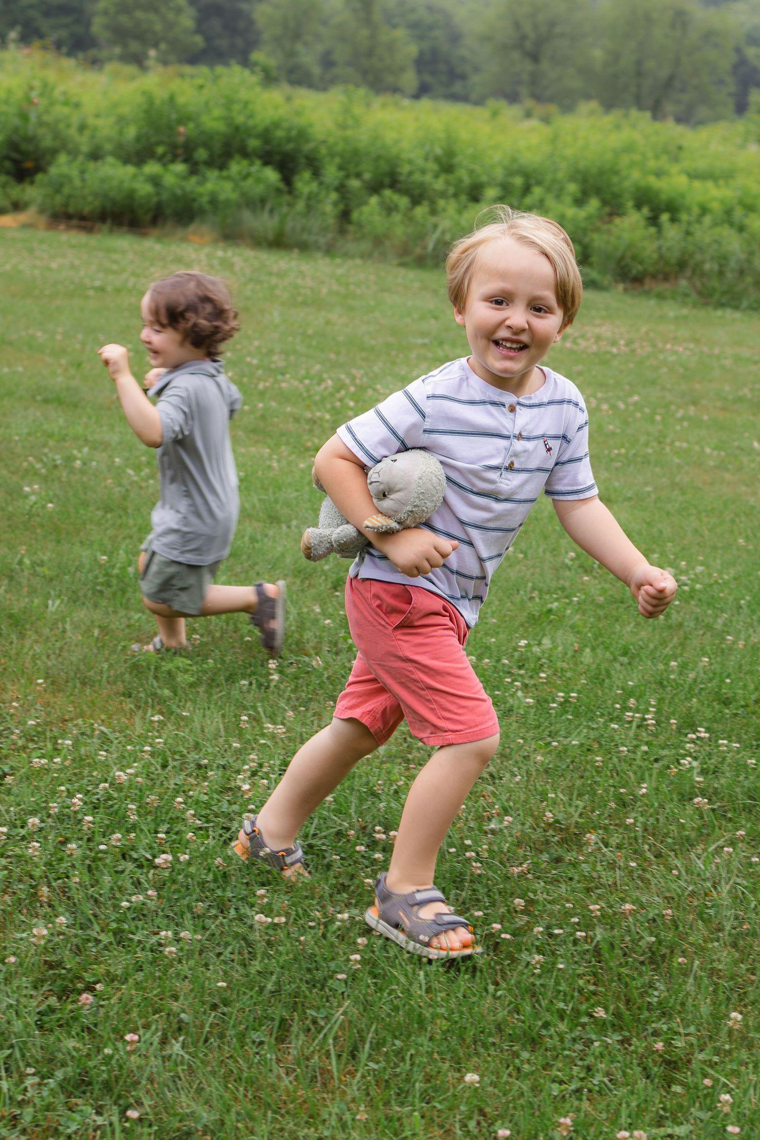 Two young boys running and laughing in a green meadow, one holding a stuffed animal.