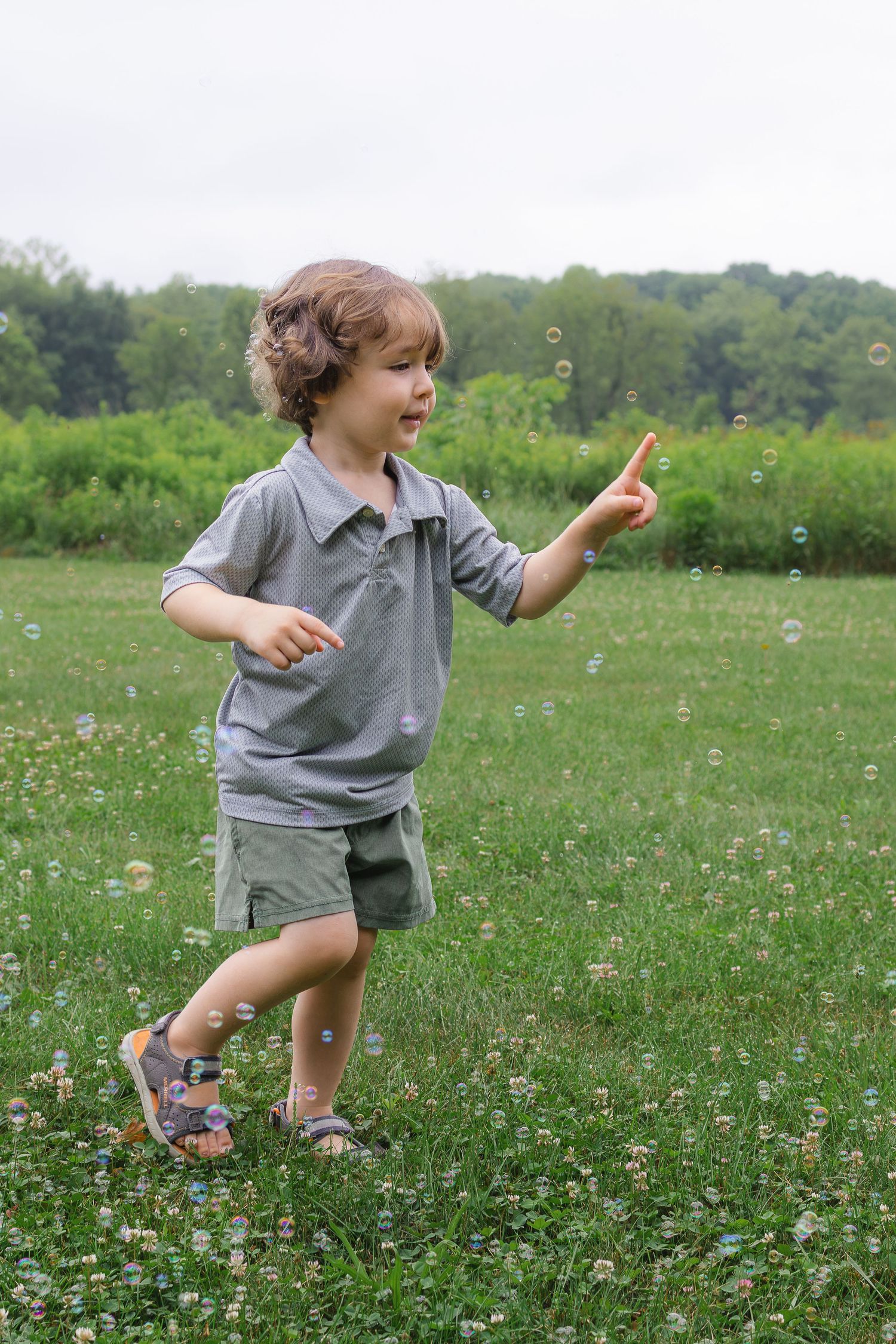Young boy in gray shirt chasing soap bubbles in a green grassy field outdoors.