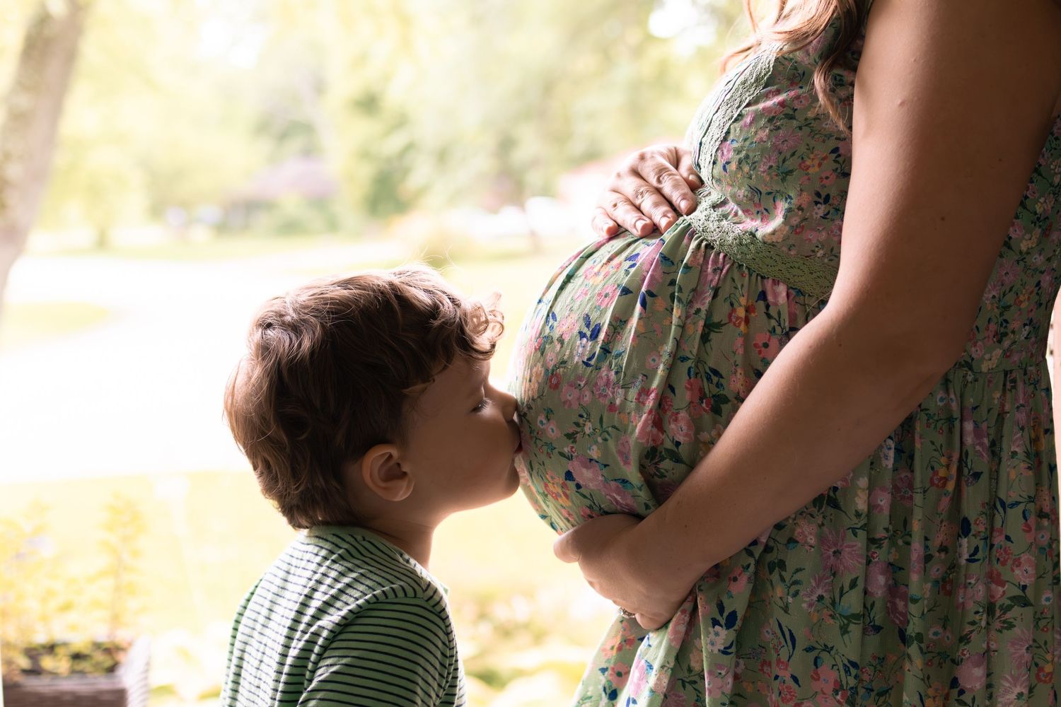 A young boy kisses his pregnant mother's belly outdoors in a sunny garden.