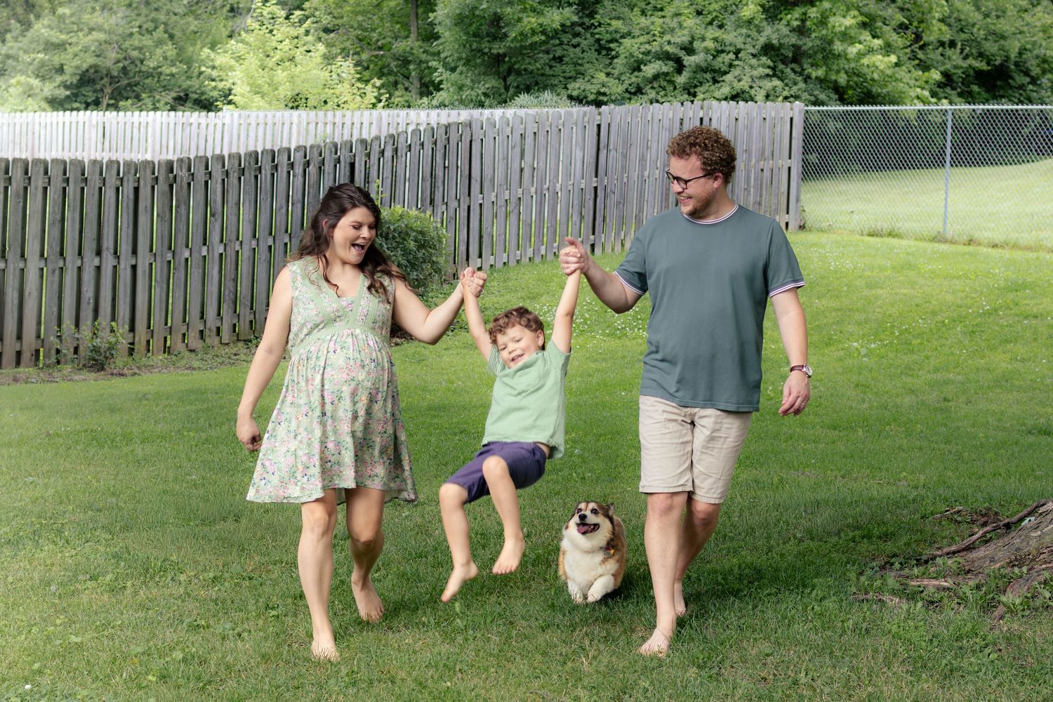 Joyful pregnant family swinging their young son by the hands while their corgi walks alongside. Mother is pregnant.
