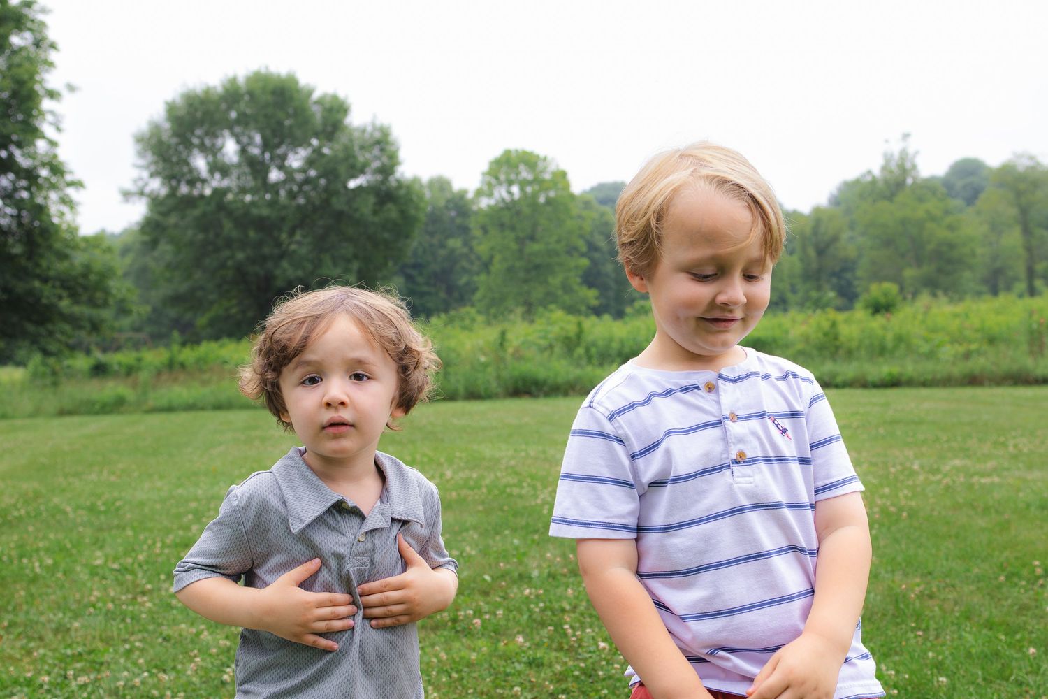 Two young toddler boys standing on green grass with trees in the background.