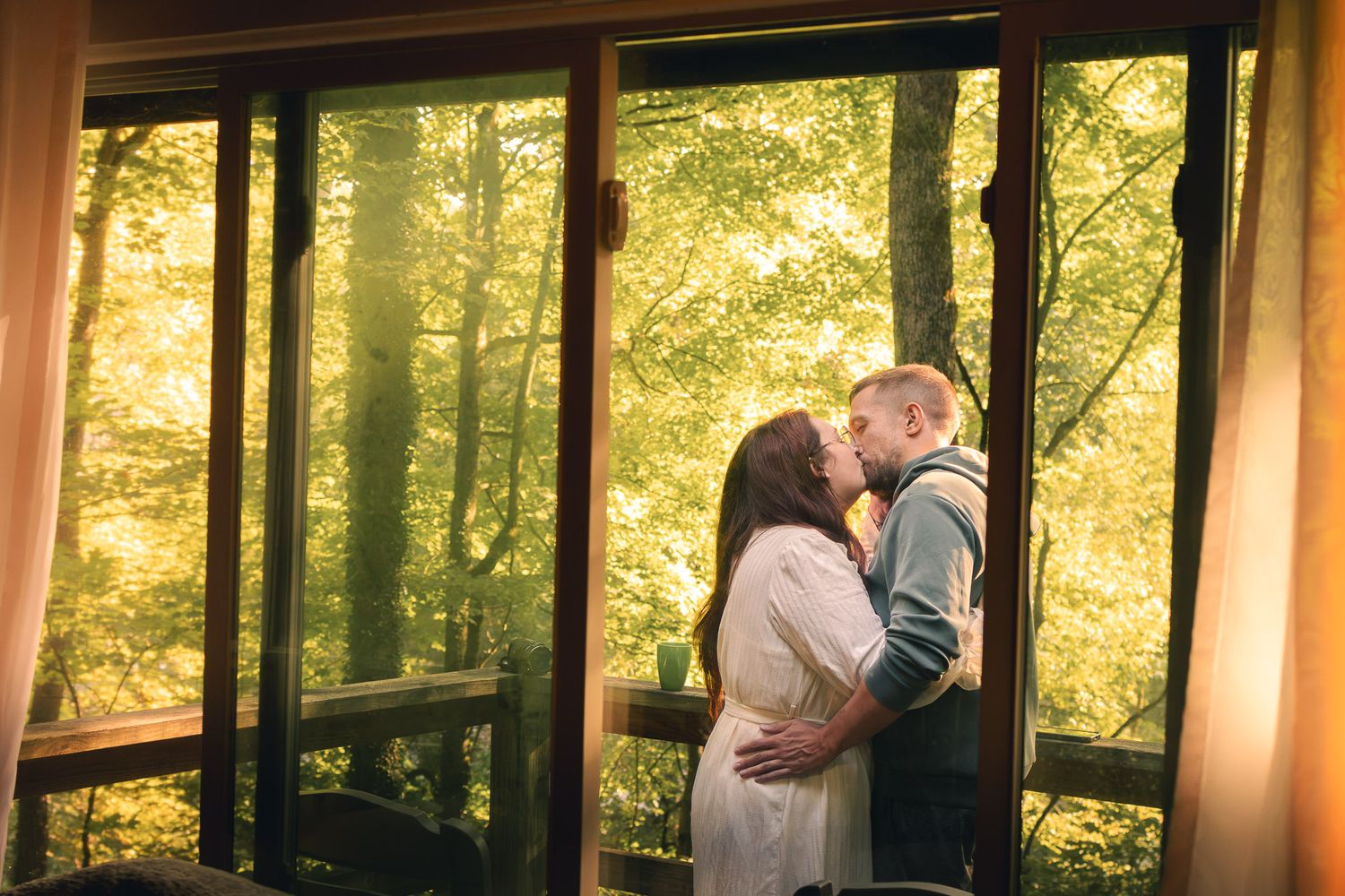 Romantic couple sharing a kiss by large windows overlooking a sunlit autumn forest cabin.