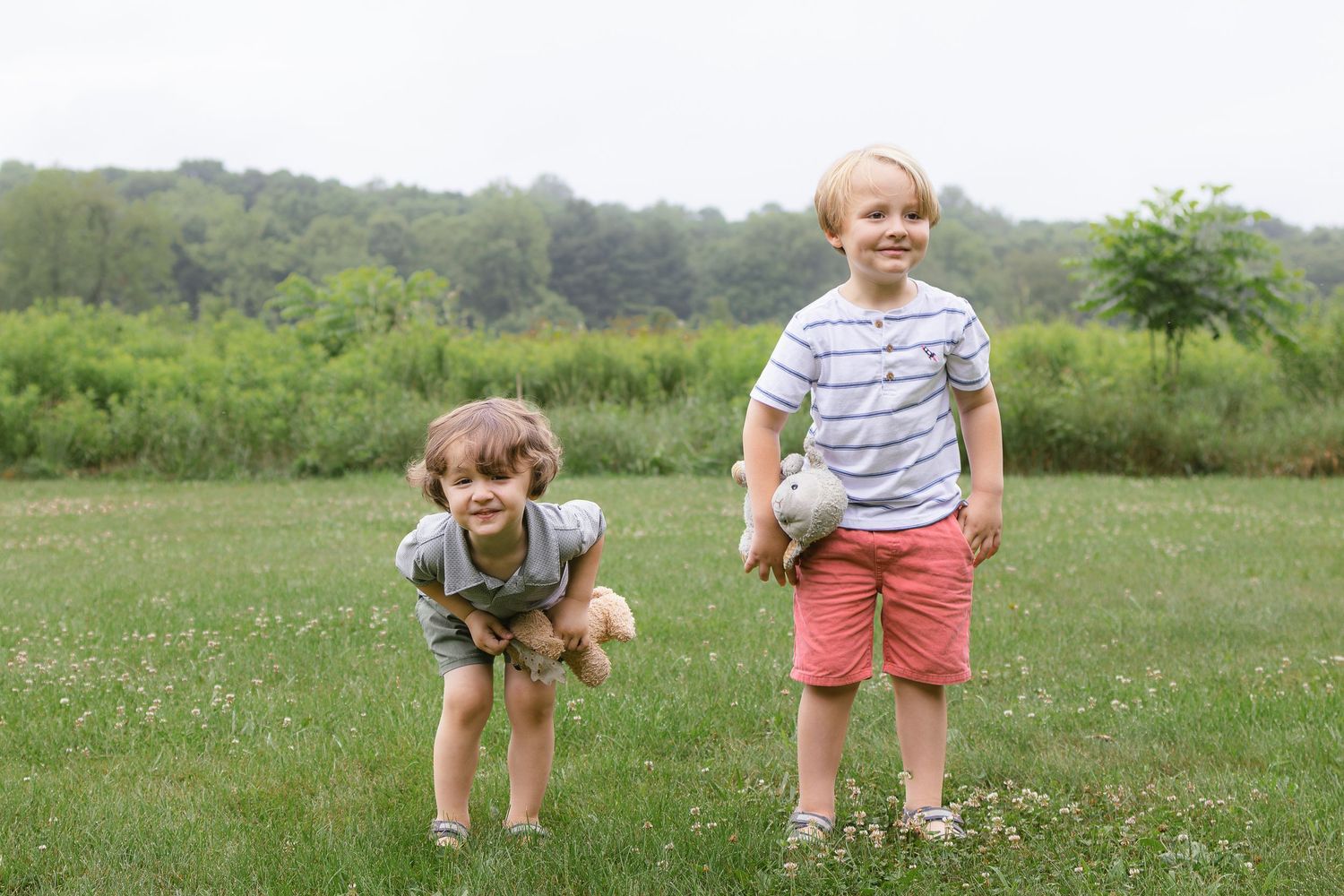 Two young boys stand and play on a green grassy field, one crouching while holding a stuffed bear.