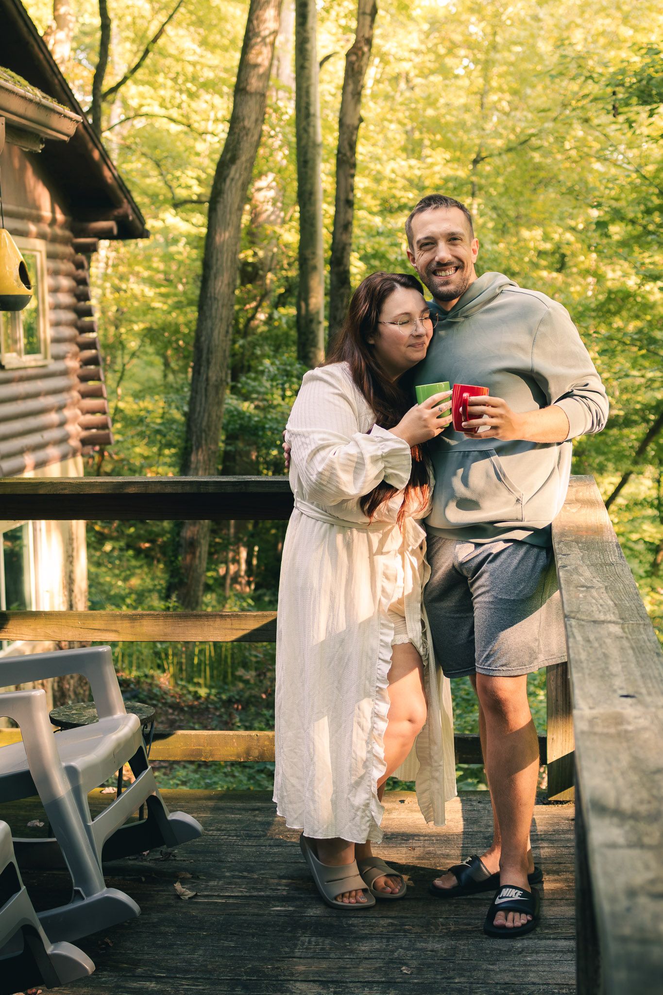 A happy couple enjoys morning coffee on a rustic log cabin deck surrounded by lush green forest trees.