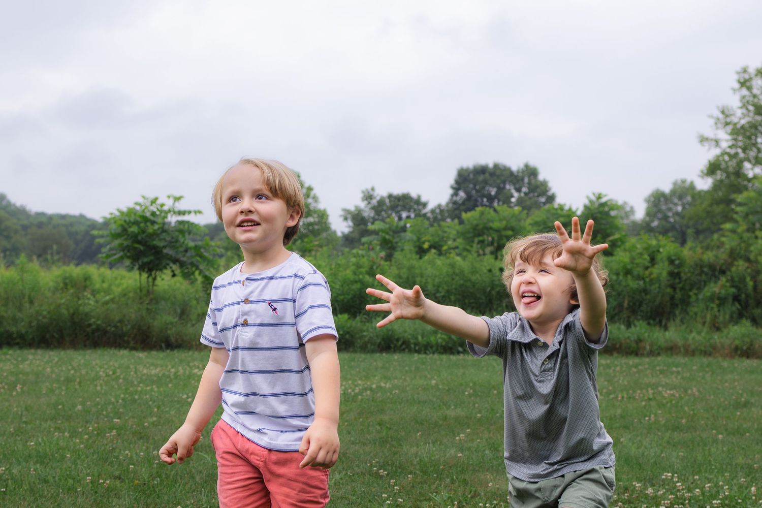 Two young boys laughing and playing together in a lush green meadow on a cloudy day.