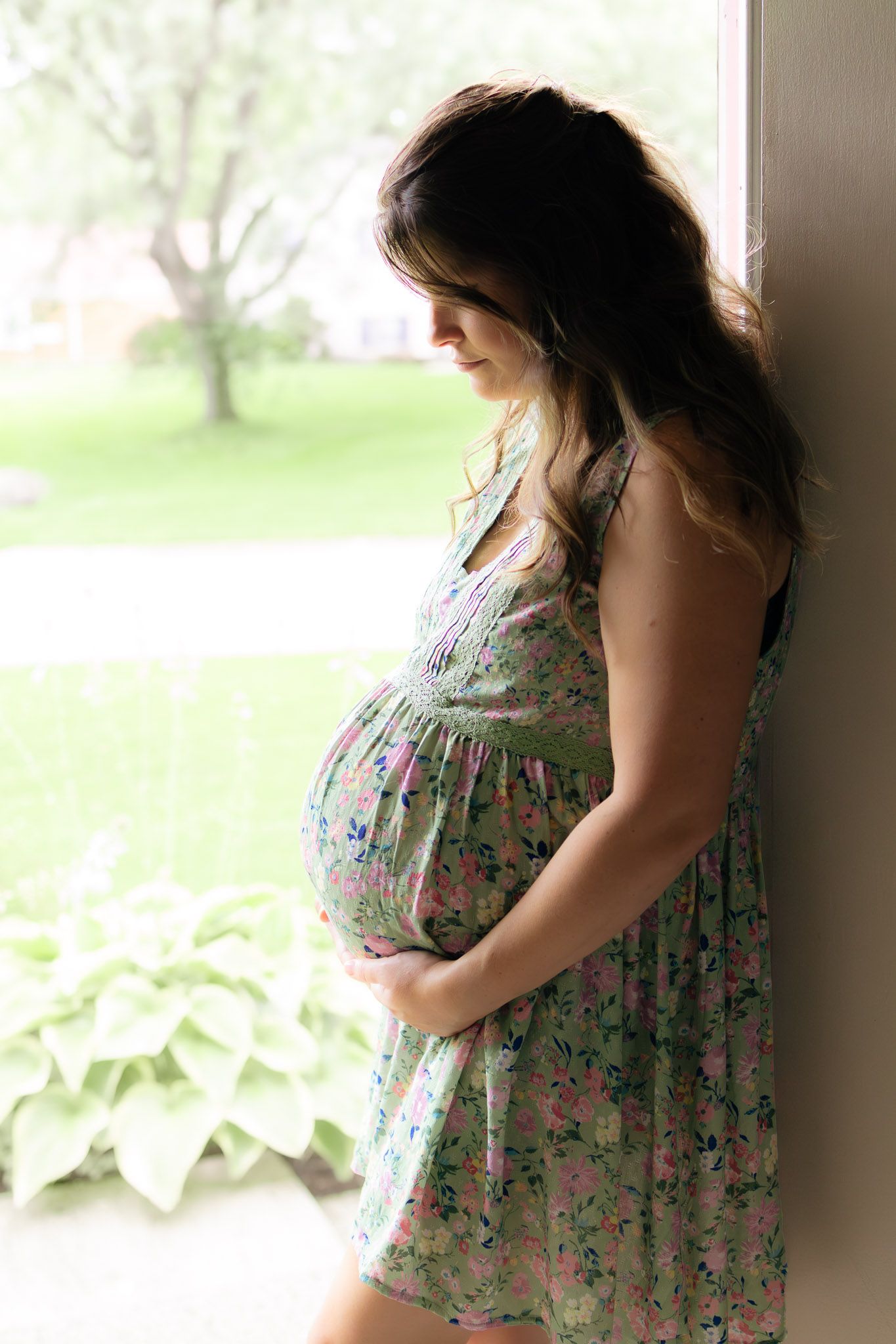 Pregnant woman in floral dress cradling her baby bump while standing by a window with green garden view. In home photo