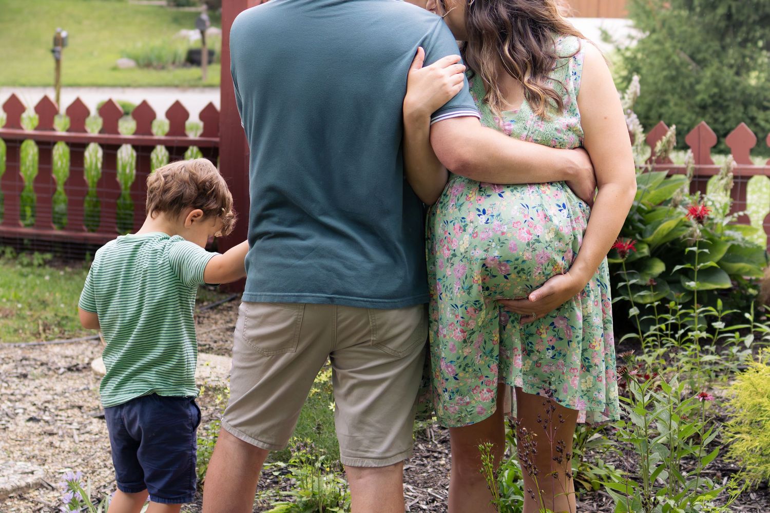 A family of three seen from behind, embracing in a lush garden near a red fence.