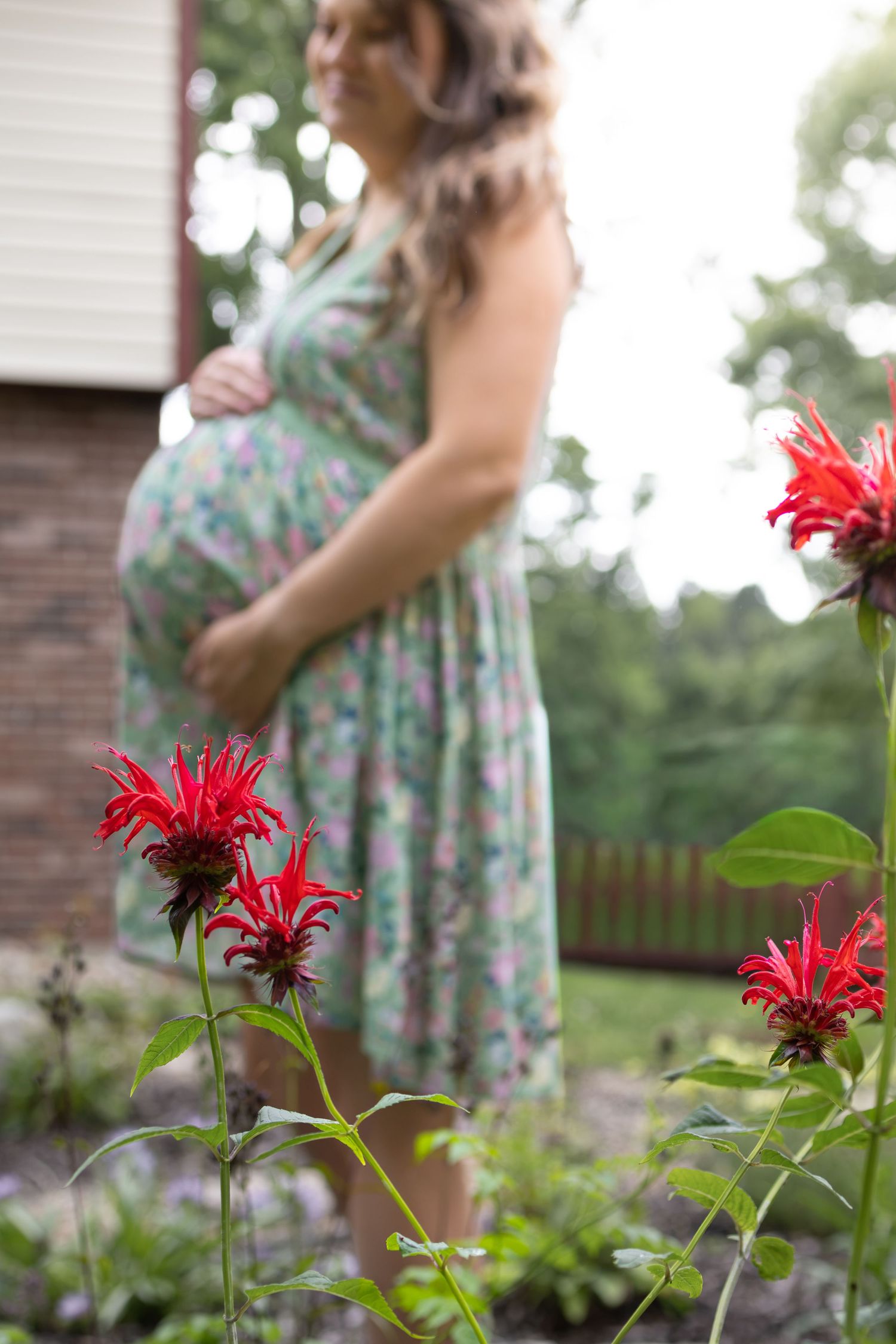 Pregnant woman in floral dress standing in garden with red flowers in foreground.