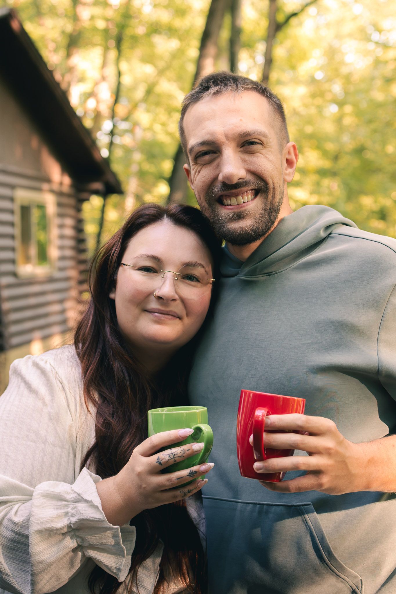 Smiling couple holding colorful mugs outdoors near a cabin in an autumn forest setting.