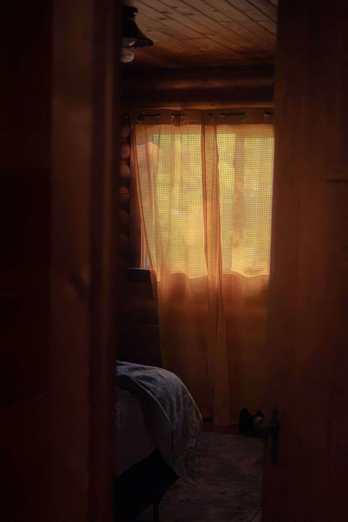 Warm golden sunlight filters through sheer curtains in a dimly lit rustic bedroom with wooden ceiling.
