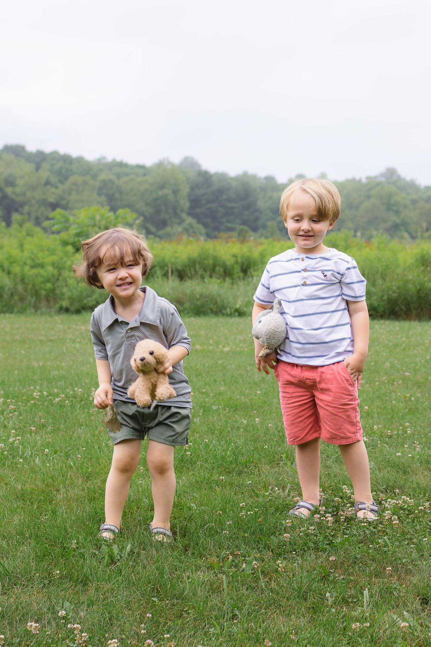 Two young boys stand on a grassy field, one laughing while holding a brown teddy bear outdoors.