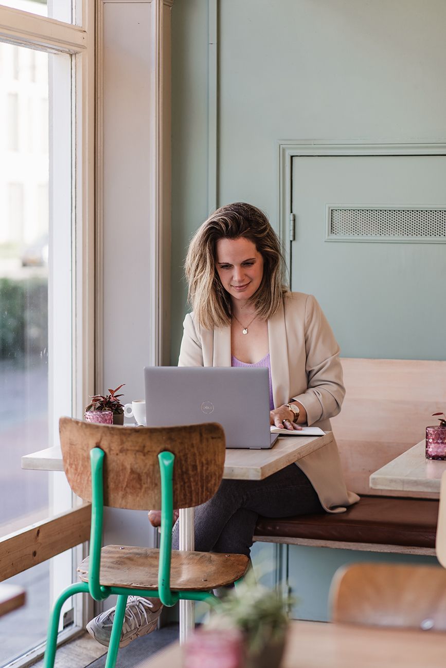 Vrouwelijke coach zit aan tafel met laptop tijdens branding shoot voor haar website.