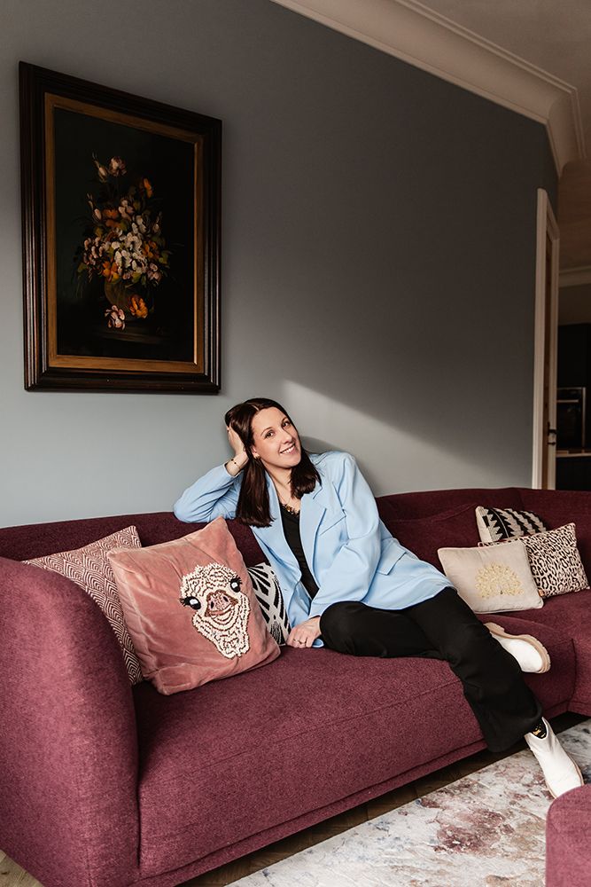 Personal branding fotografie voor een interieurontwerper met een warme sfeer bij haar kleurrijke huis. Ze zit op een bordeaux rode bank.