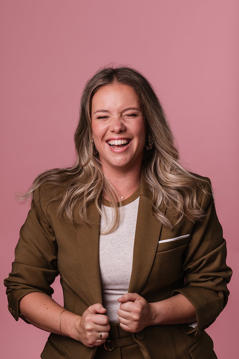 Zakelijke kleurrijke portretfotoshoot met een vrouwelijke ondernemer die lachend op de foto staat en met roze achtergrond.