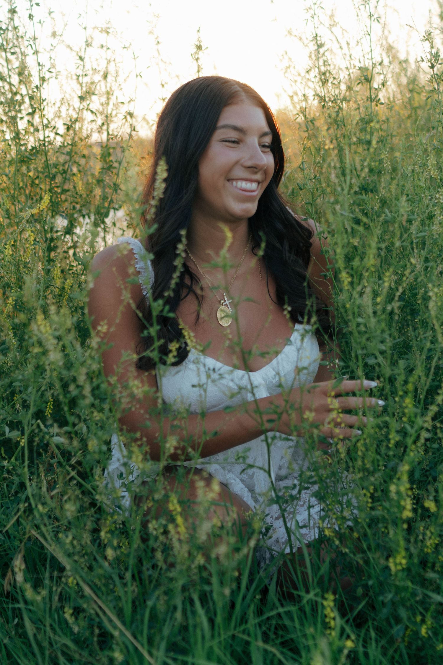 Smiling woman with dark hair in white dress surrounded by tall green wildflowers at sunset.