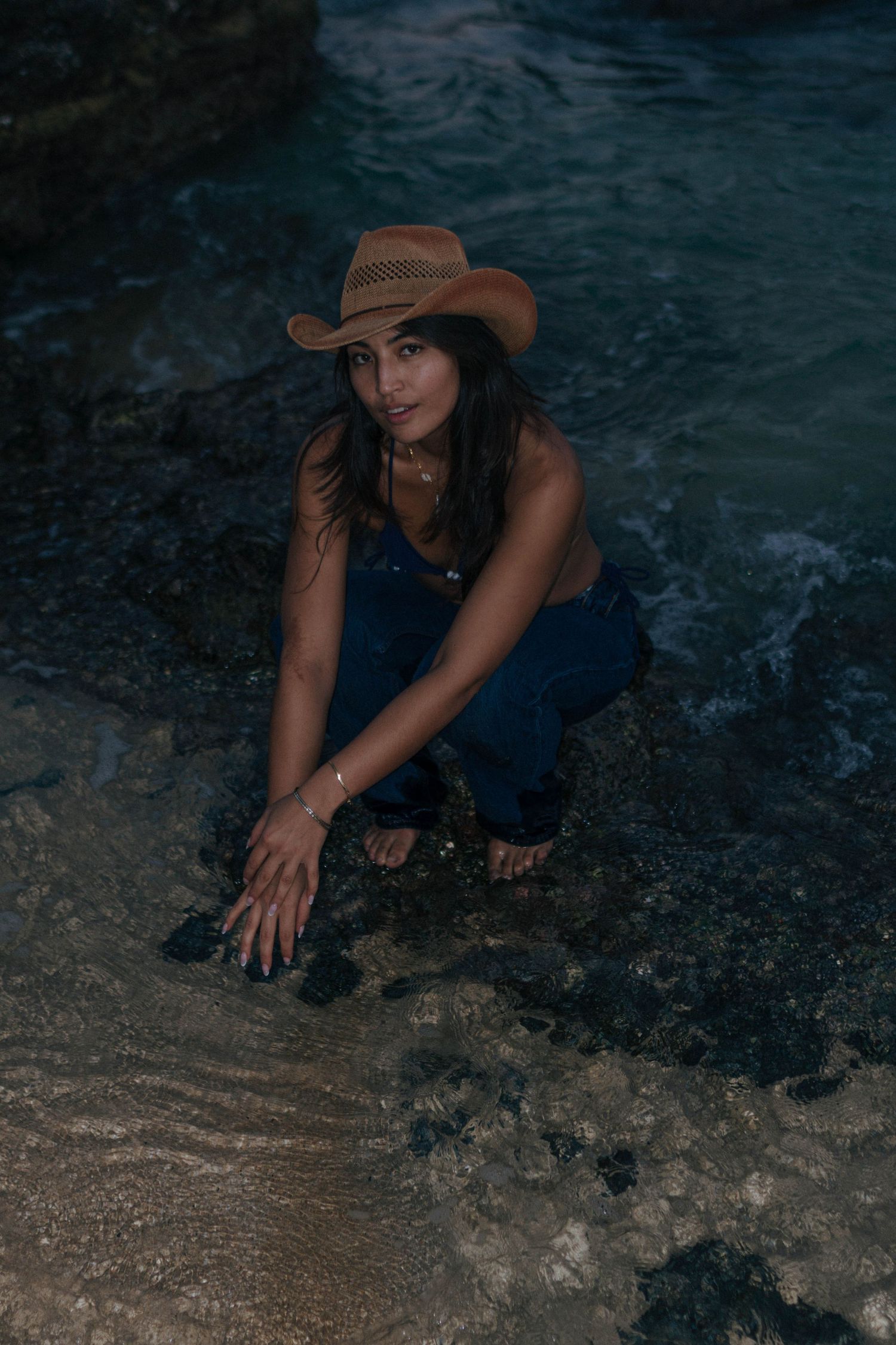Woman in denim outfit and cowboy hat crouches near ocean water on rocky beach at night.