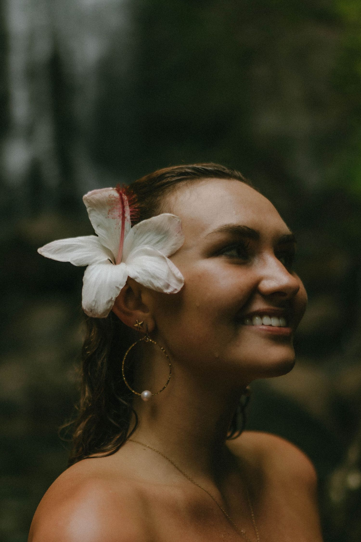Smiling woman with white hibiscus flower in wet hair wearing hoop earrings near waterfall.