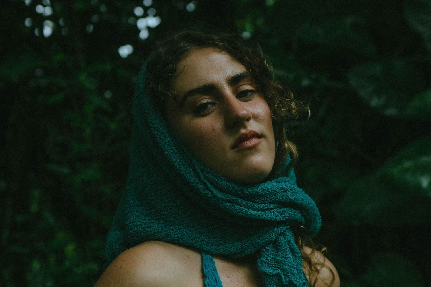 Young woman with teal scarf draped over head gazes upward surrounded by dark green foliage.