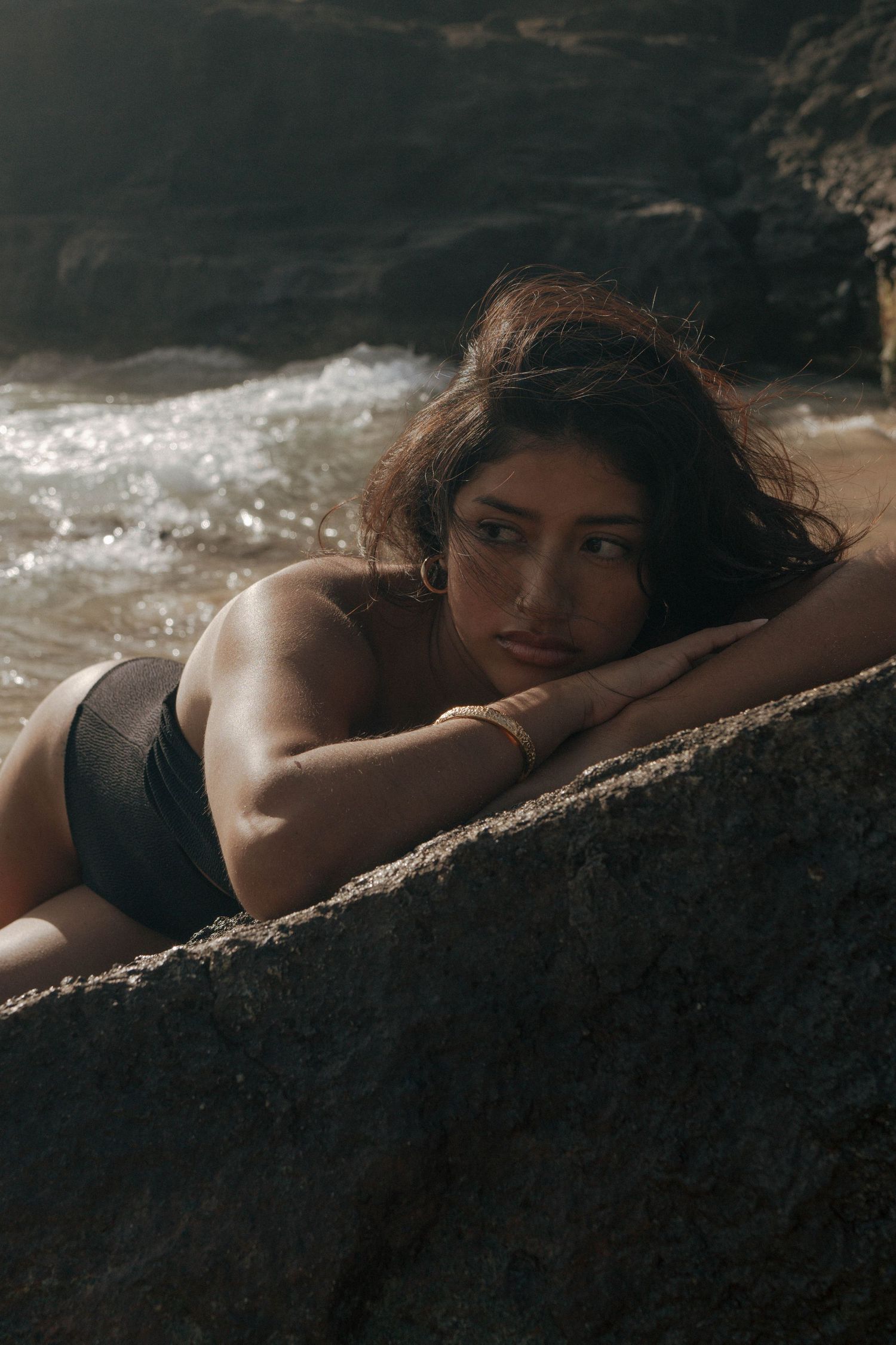 Woman in black bikini lying on rocky beach with ocean waves in background.