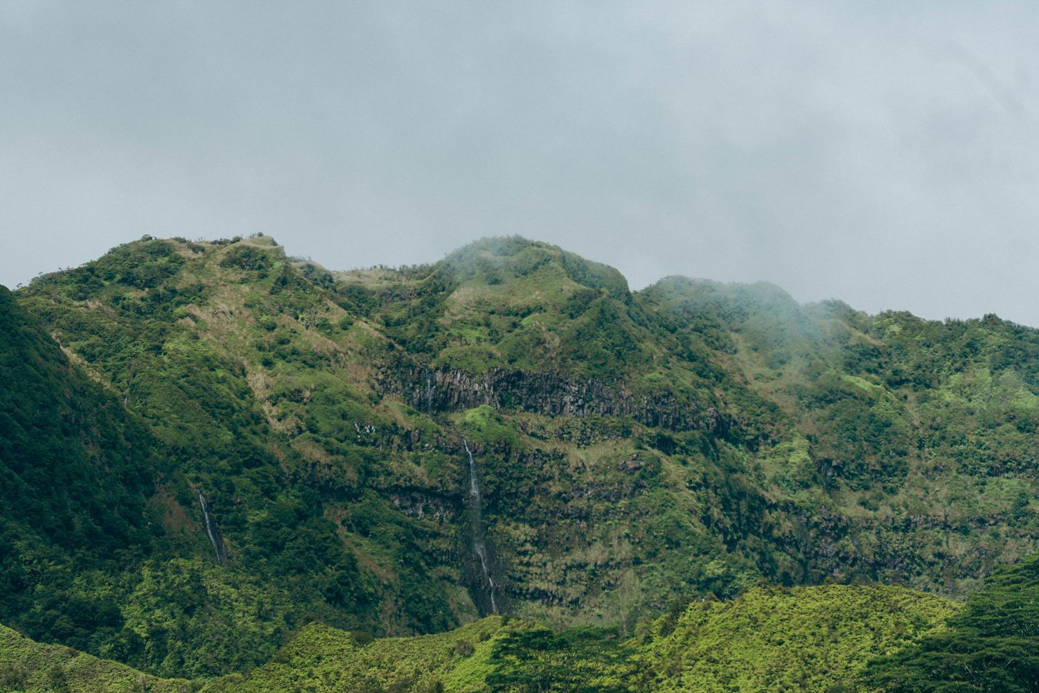 Lush green tropical mountain with cascading waterfall under misty cloudy sky.
