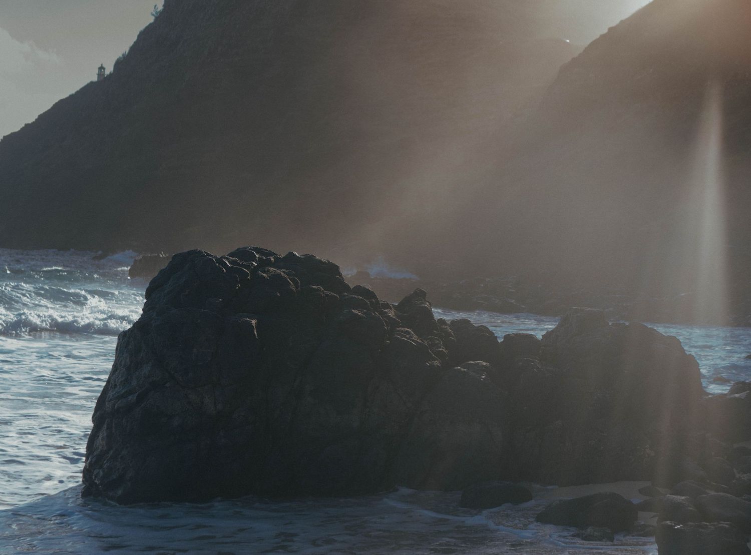 Golden sunlight rays bursting over rugged coastal rocks with crashing ocean waves below.