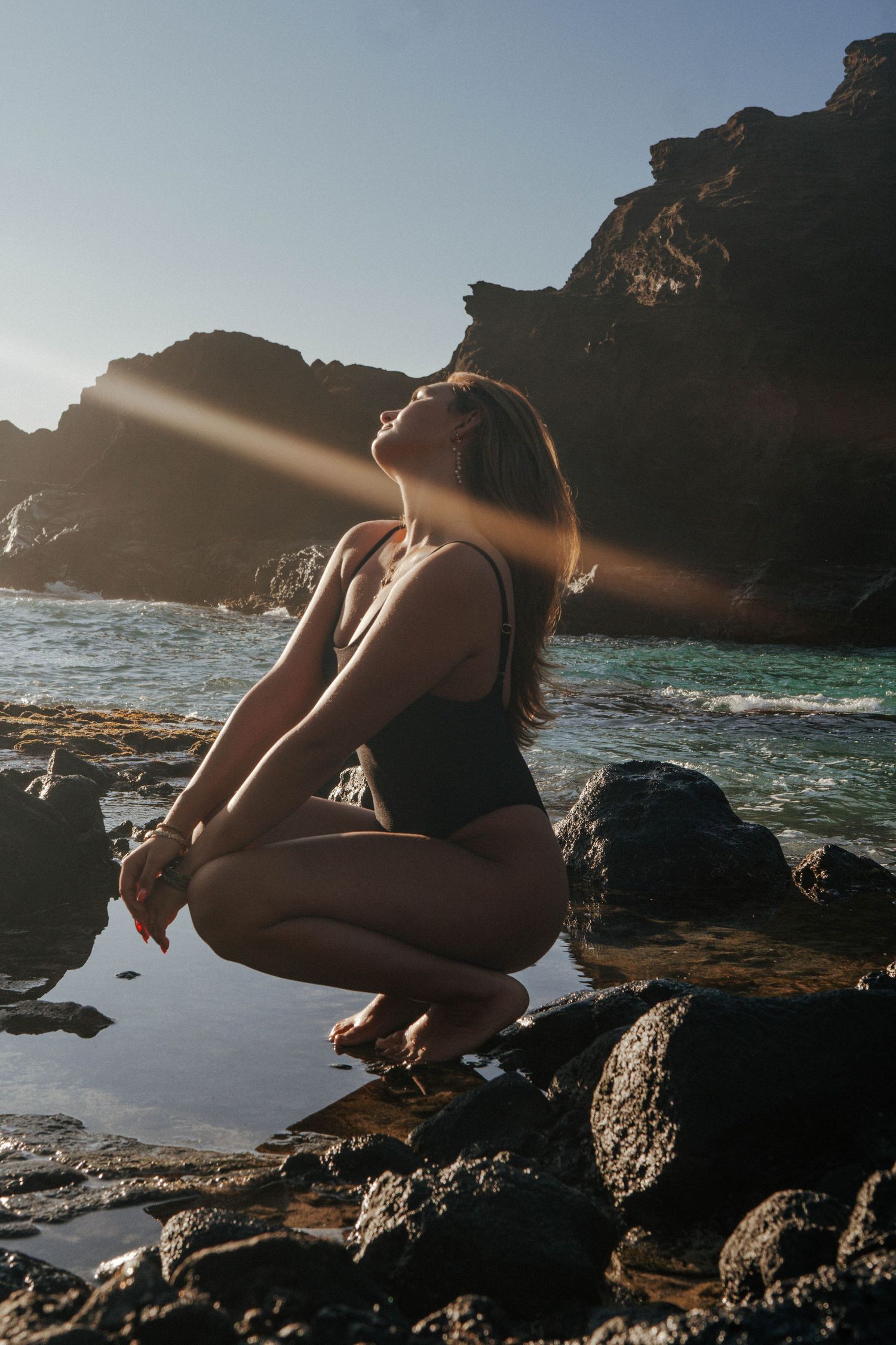 Woman in black swimsuit crouching on rocks with sunlight rays by ocean.