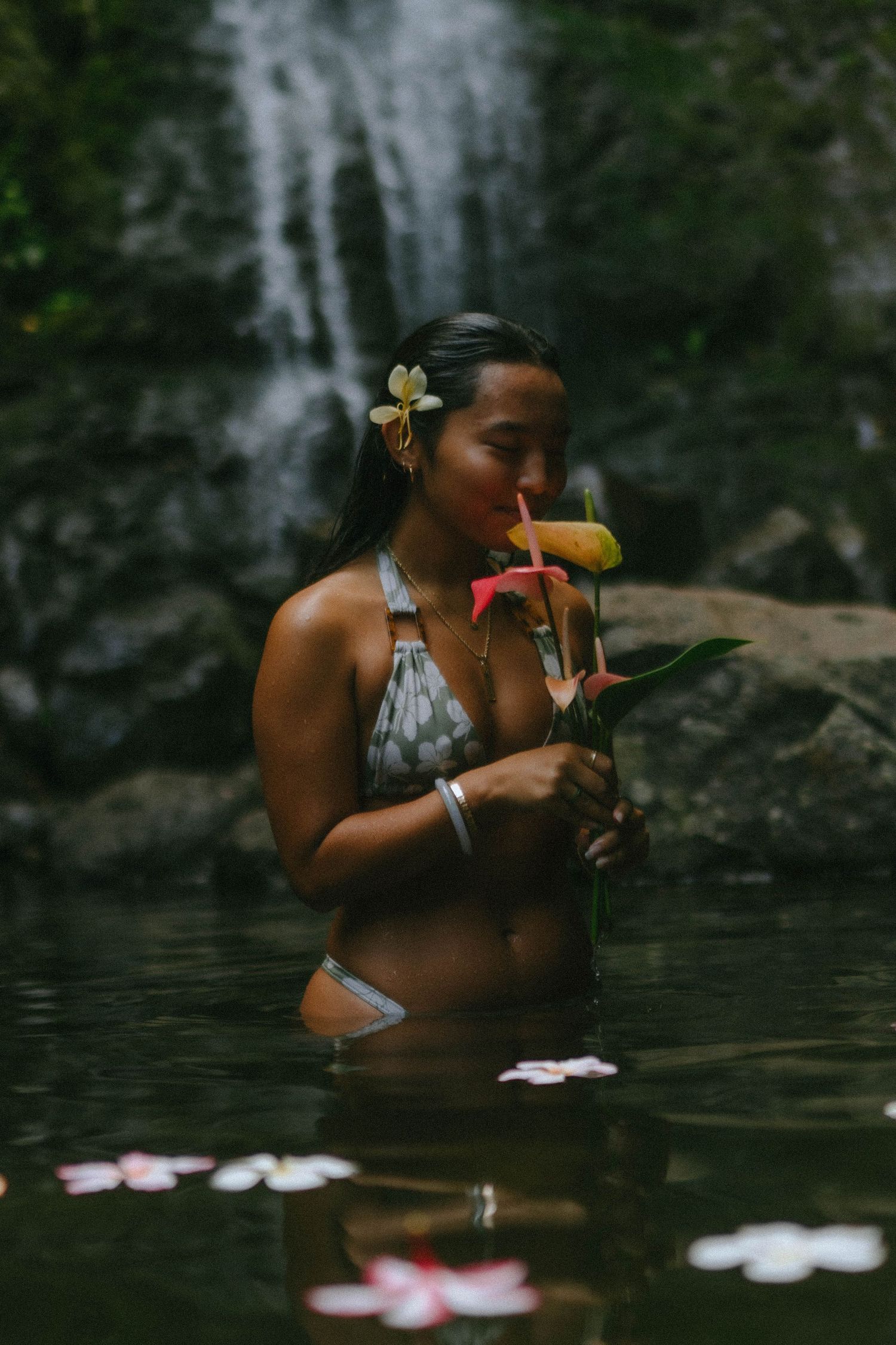 Woman in gray bikini holds tropical flowers near her face while standing in waterfall pool.