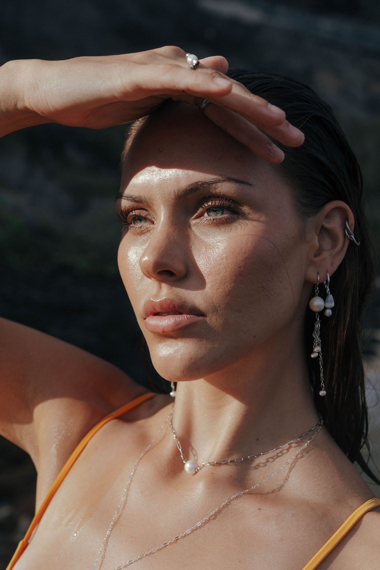 Close-up portrait of woman with wet skin shielding eyes from sun wearing pearl earrings and necklace.