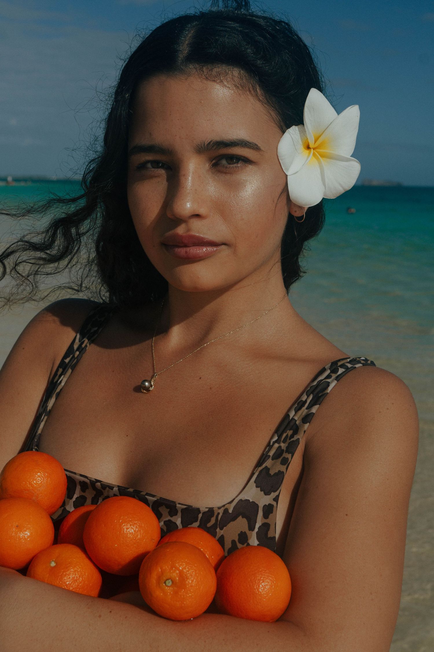 Woman with white plumeria flower in hair holding oranges wearing leopard print swimsuit at beach.