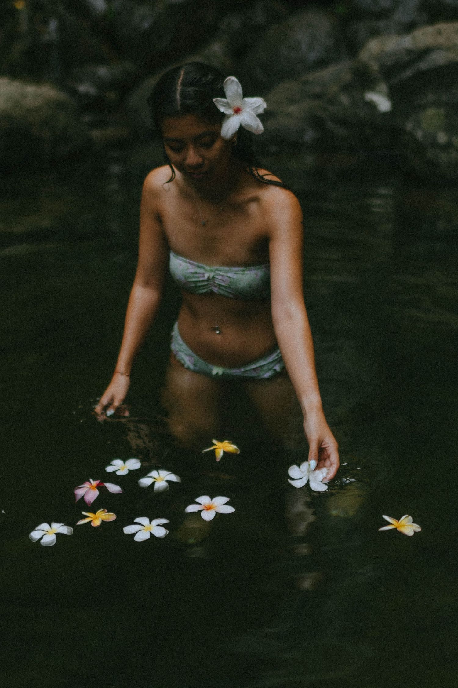 Woman in sage green bikini arranges floating plumeria flowers in dark tranquil waterfall pool.